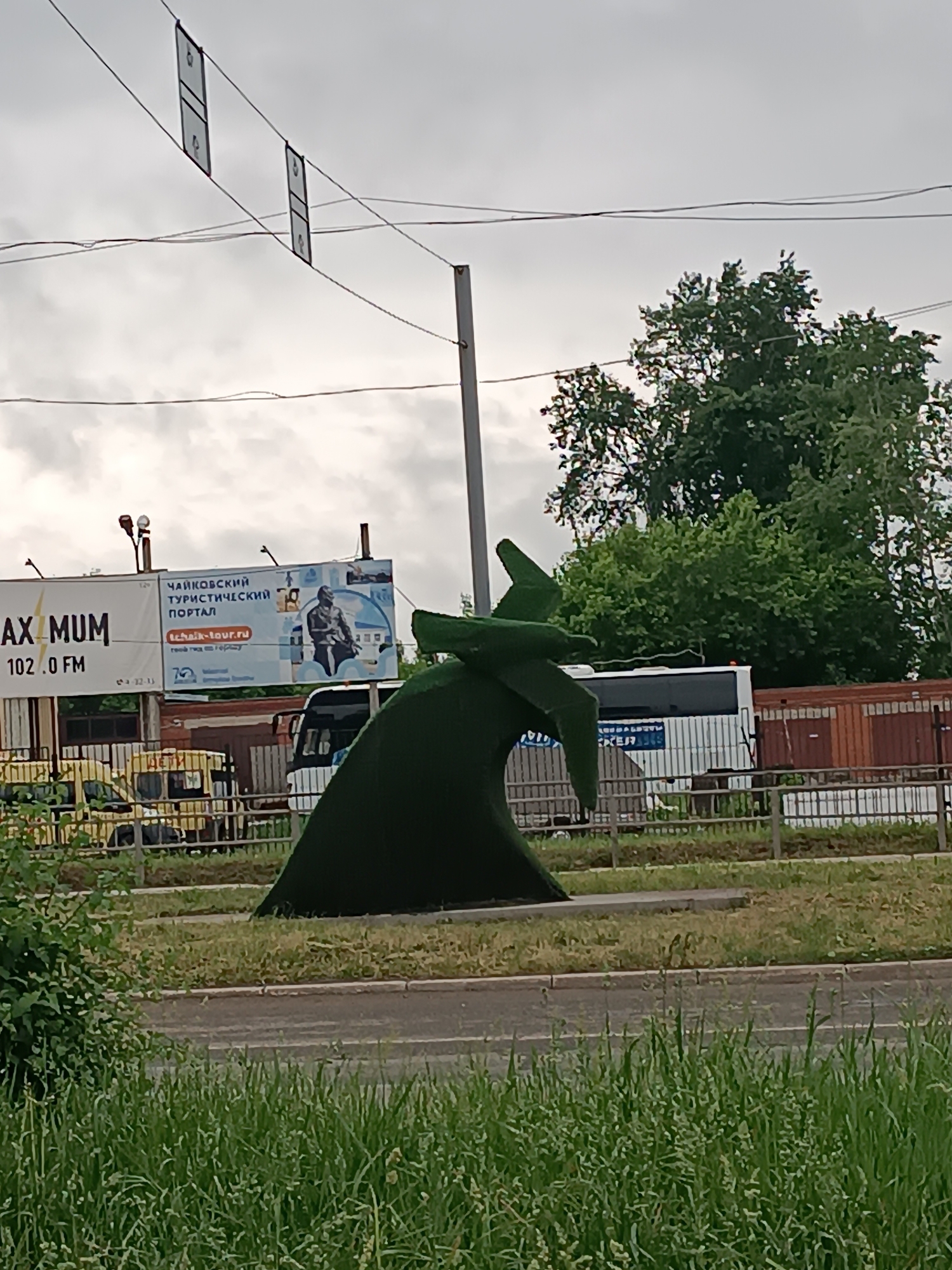 The image shows a green sculpture of an airplane placed on a grassy area in Chaikovsky, Russia. Behind the sculpture, there is a white bus and a billboard advertising the "Chaikovsky Tourist Portal." The billboard also displays an advertisement for a radio station, "Maximum 102.0 FM." A metal fence separates the grassy area from the road. In the foreground, there is tall green grass growing along the roadside. Overhead, there are utility lines and rectangular signifiers that seem to be suspended on the lines. Trees are visible in the background.