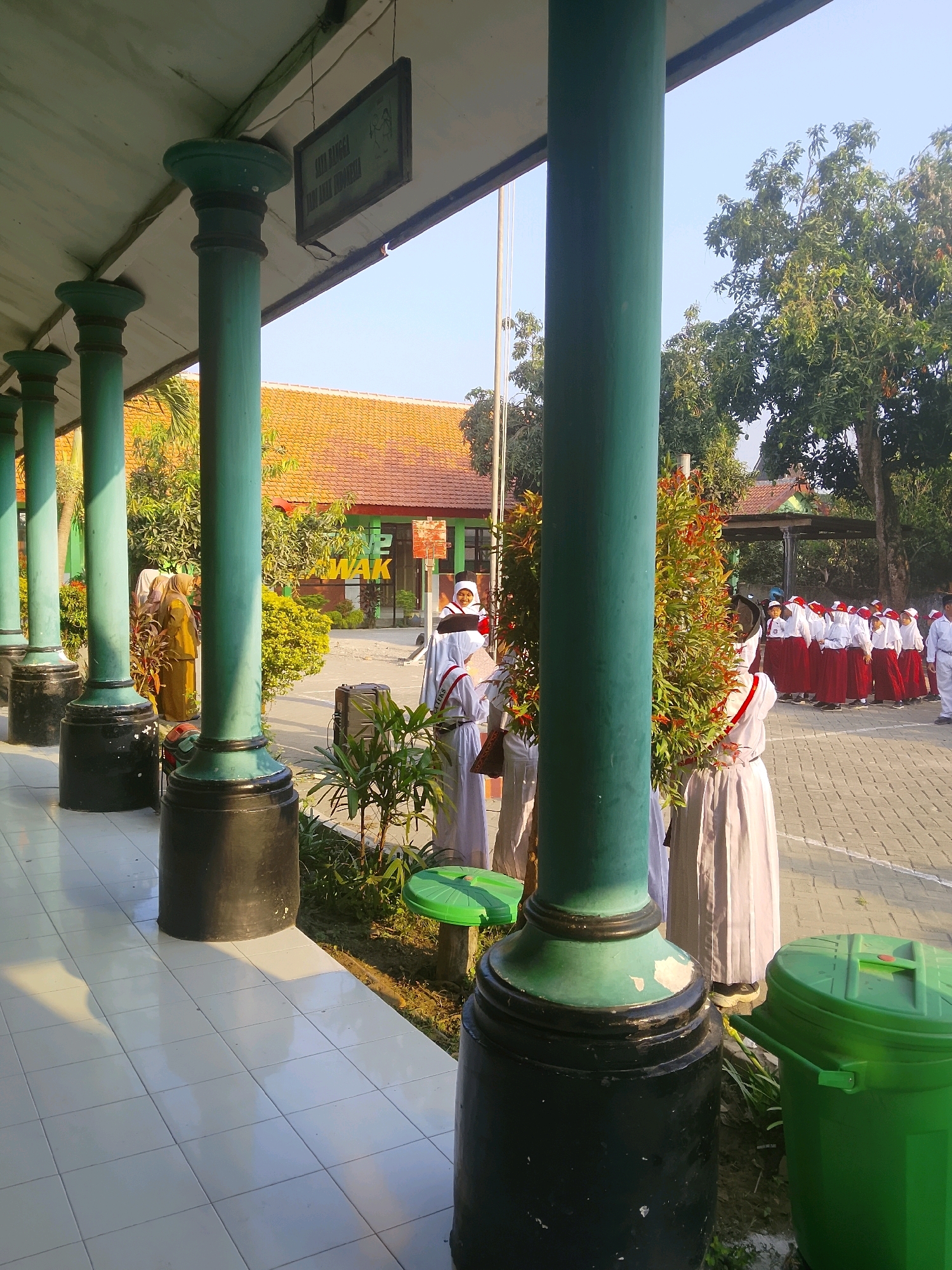 The image shows an outdoor corridor of a school building in Kepuh, Indonesia. The corridor is supported by several large green pillars with black bases. The floor is tiled with white square tiles. In the background, there is a building with an orange tiled roof and some trees. A group of children in uniform, likely students, are standing in an open area outside the corridor. Some of them are playing music with drums. The sky is clear and blue. A green trash can is visible near one of the pillars. There is a framed poster hanging from the ceiling of the corridor.