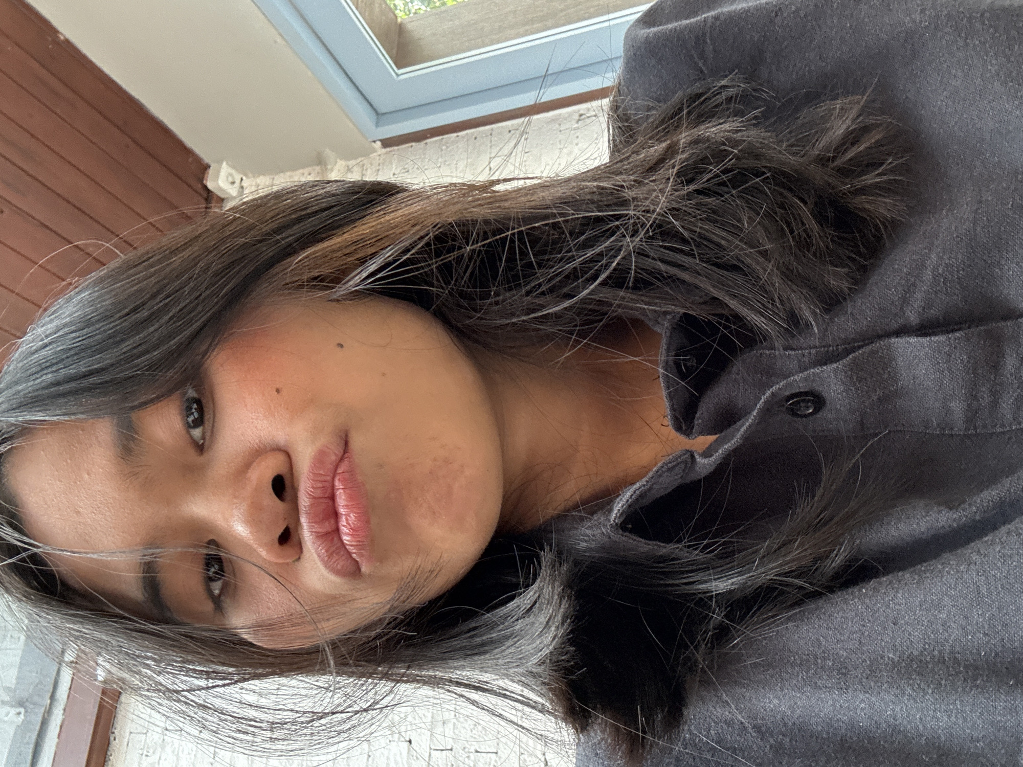 A selfie taken from a low angle shows a young woman with dark hair and brown eyes puckering her lips. She is wearing a dark gray collared shirt. In the background, a white brick wall, a wooden ceiling, and a blue window frame are visible. The scene appears to be outdoors in a residential or commercial area of South Jakarta, Indonesia.