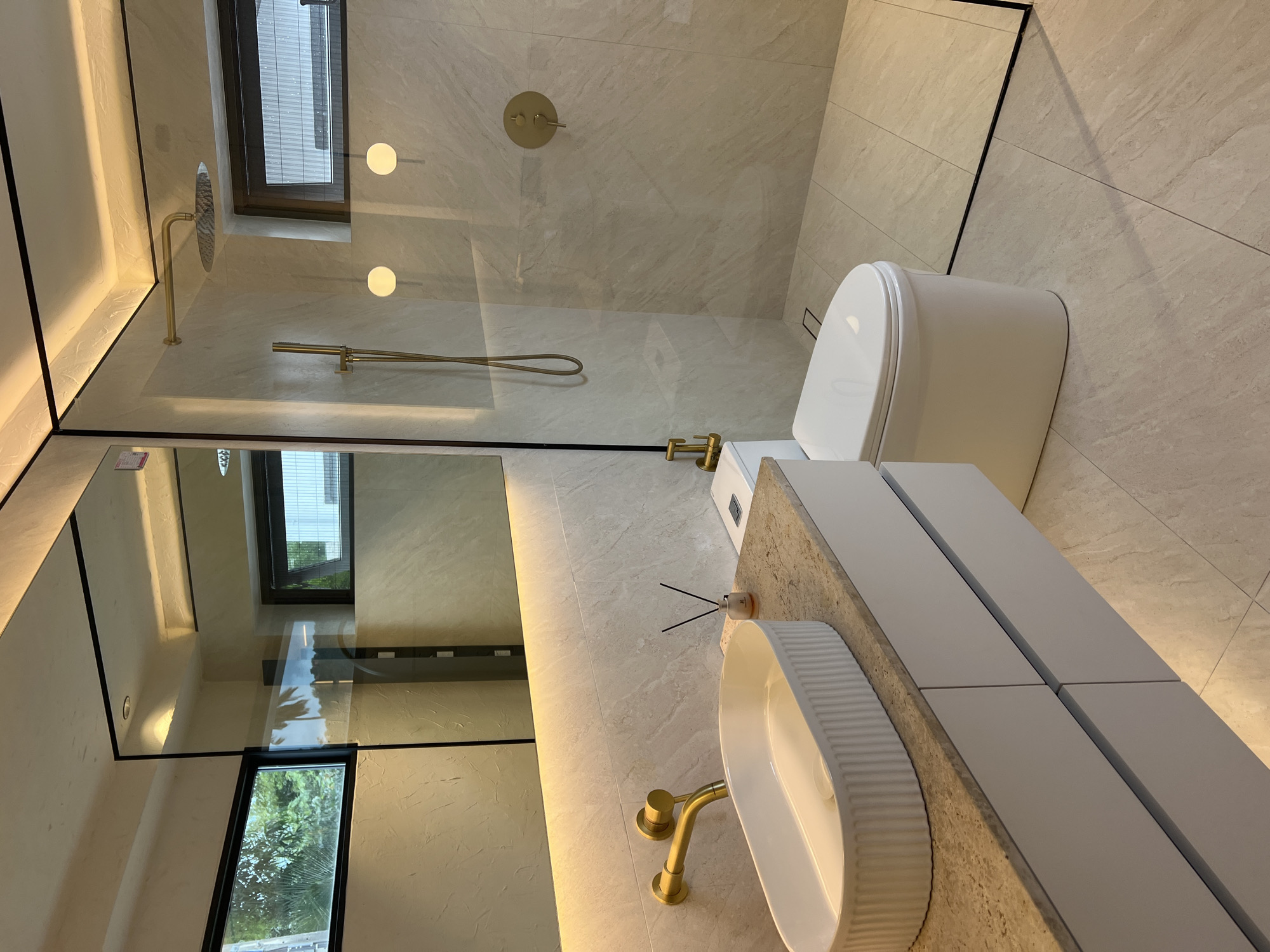 A high-angle shot depicts a modern bathroom setting, appearing bright and clean, in what could be a residence or hotel in the Baan Bang Rak area of Thailand. The room features a toilet with a closed lid, a sink with a white, ribbed exterior, and a vanity with a light-colored countertop and white, block-style cabinetry. Gold fixtures are notable for the faucet and shower components. The shower area is enclosed by a glass partition. Light-colored tiles cover the floor and walls. A mirror reflects a window, potentially revealing an outside view, possibly of greenery. The overall design indicates a focus on modern aesthetics and functional use of space. There are no people visible in the frame, and the scene is static. A reed diffuser sits next to the sink.
