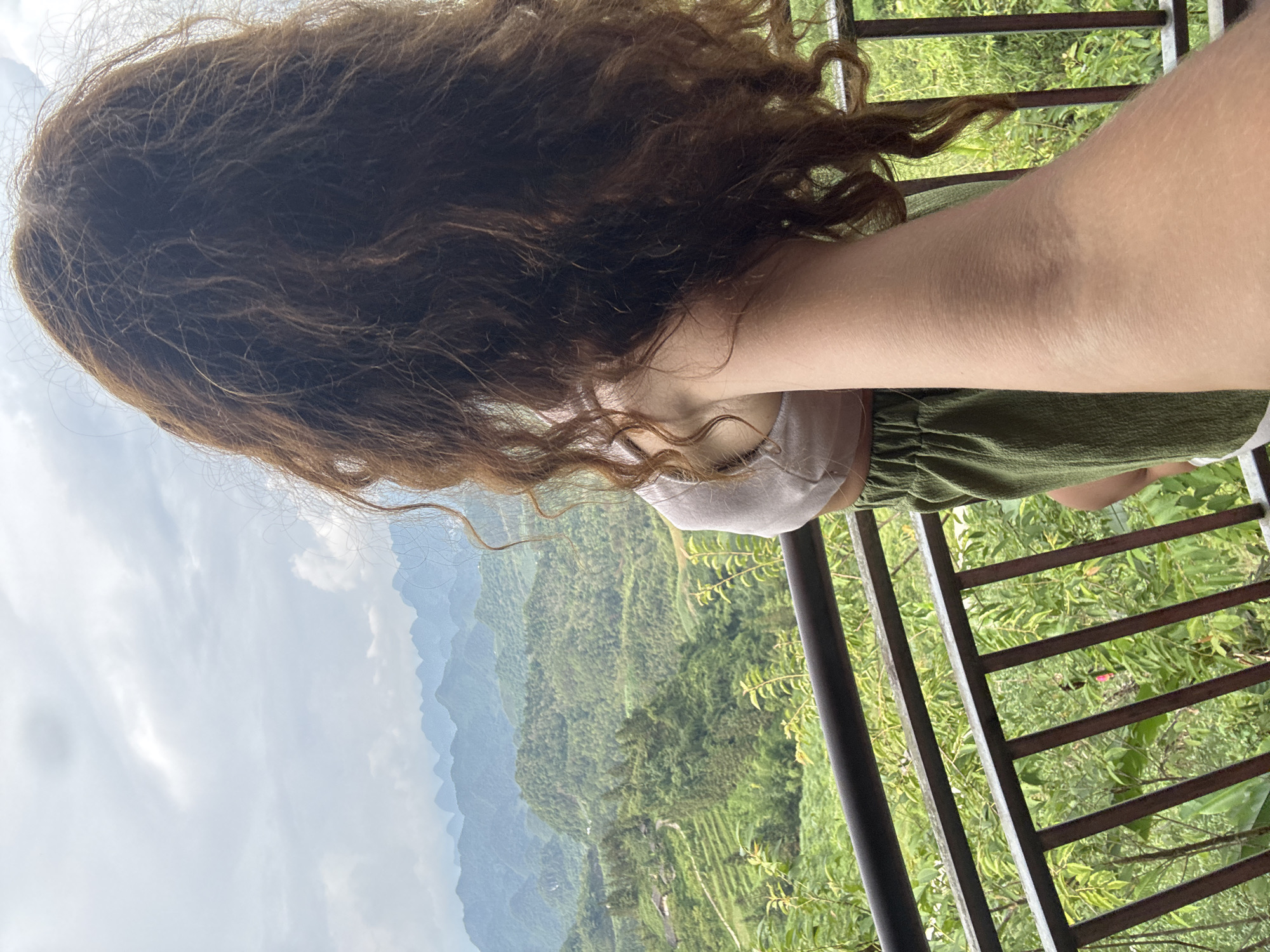 A woman stands on a balcony overlooking a lush, green valley in what appears to be the Quản Bạ Commune area of Vietnam. Her dark, curly hair is prominent in the foreground. She is wearing a light-colored top and green shorts. A dark metal railing lines the edge of the balcony, providing a view of the terraced hills and mountains in the background. The sky is overcast, creating a diffused light across the scene.