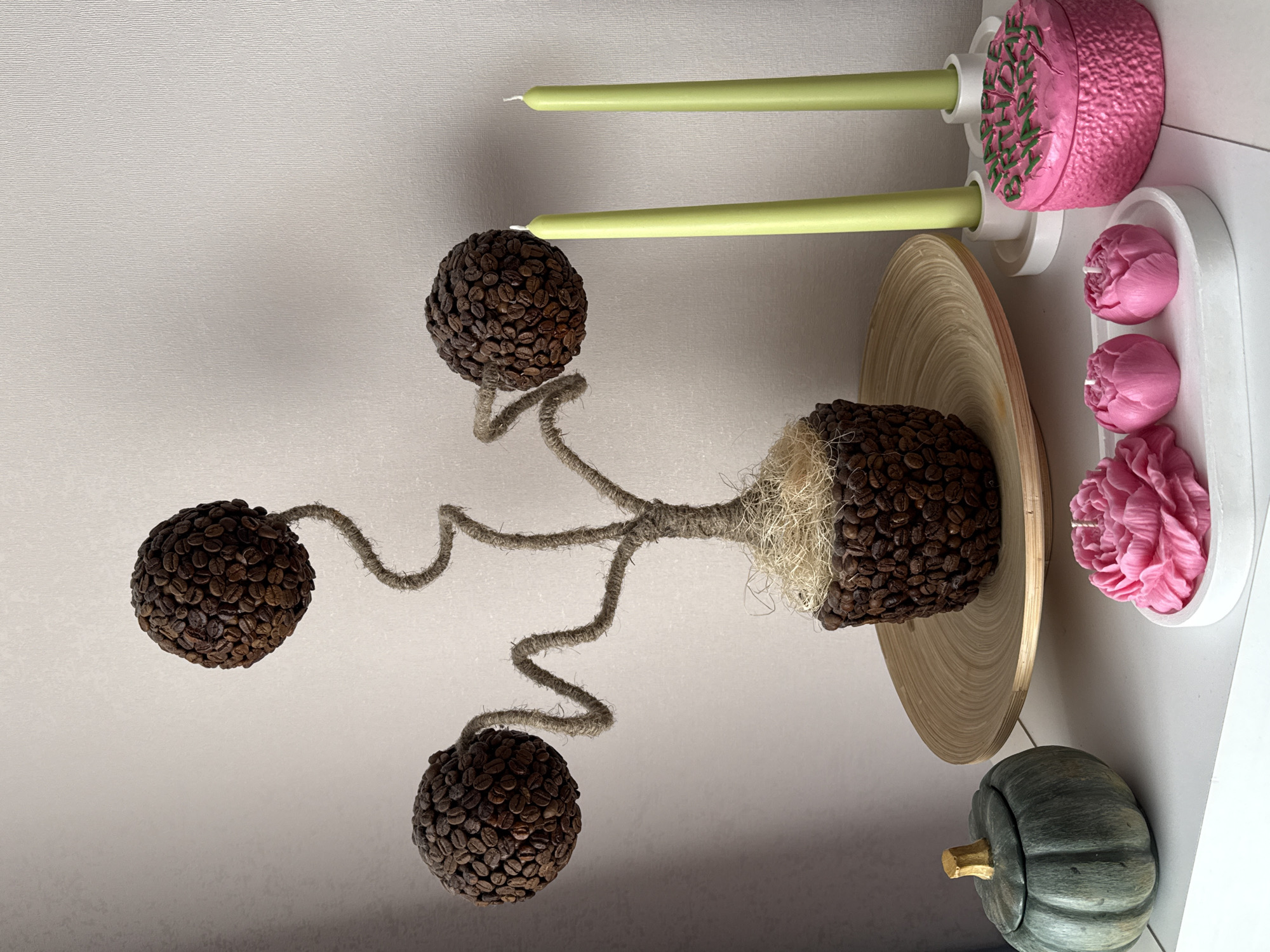 On a light-colored surface in what appears to be an interior setting, there is a decorative arrangement featuring a tree-like structure made of twine and coffee beans. The structure has a central "trunk" and three spherical "branches" covered in coffee beans. It sits on top of a wooden plate. Beside the plate, there is a white tray containing three pink flower-shaped objects, likely candles or soaps. Adjacent to the tray is a pink, textured candle holder with two green candles. Near the bottom of the frame, there is a small, gray-green pumpkin decoration. The location is not discernible from the image and cannot be confirmed as Krasnodar, Russia.