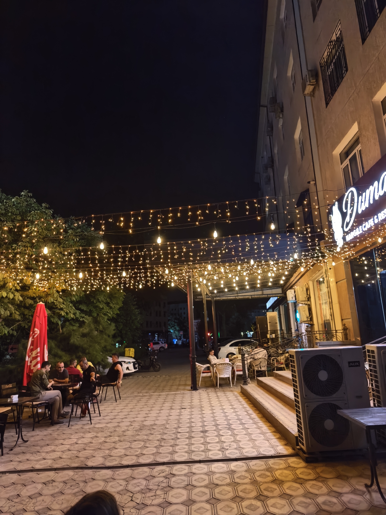The image captures a nighttime scene outside a cafe in Fergana City, Uzbekistan. A row of tables and chairs are set up on a patterned paved area, illuminated by strings of warm-toned fairy lights suspended overhead. Several people are seated at tables, engaged in conversation or dining.
To the right, the facade of a building is visible, featuring a sign with the name "Duman" in a stylized font, indicating it is a cafe and restaurant. The building appears to be an apartment block or a commercial building with multiple floors and windows. Several air conditioning units are mounted on the exterior wall near the entrance, one of which is clearly branded "AUX".
In the background, trees with dense foliage are illuminated by the fairy lights. A red umbrella is prominent near the outdoor seating area. The street beyond the cafe shows some traffic, with cars and a scooter visible. The overall atmosphere is that of a vibrant outdoor dining and social space at night.