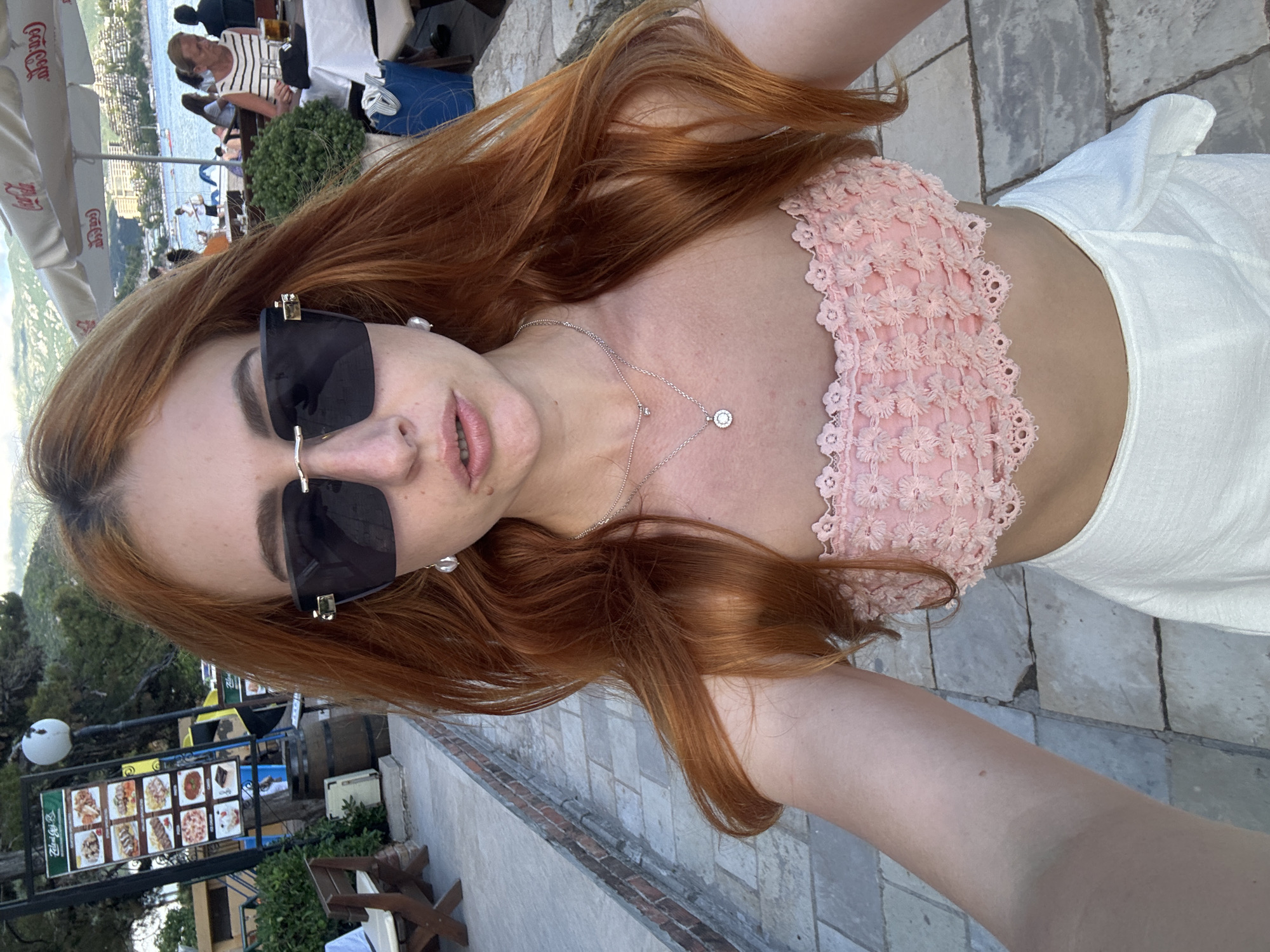 A woman takes a close-up selfie in an outdoor setting, likely a restaurant or cafe, with a scenic coastal view. She has long, reddish-brown hair and is wearing large, dark square-framed sunglasses with subtle gold embellishments. Her lips are slightly parted in a neutral expression. She is adorned with a delicate silver necklace featuring a small circular pendant and a matching pair of pearl-like earrings. Her attire consists of a light pink strapless top with intricate floral lace or crochet detailing and a white, textured skirt.
The background reveals a vibrant outdoor atmosphere under bright daylight, possibly late afternoon. Behind the woman, several white umbrellas, some branded with "Coca-Cola," provide shade for patrons seated at tables. In the mid-ground, a woman with a striped top is visible among other indistinct figures. Beyond the establishment, a calm body of water, likely the sea or a bay, stretches out with boats dotting its surface, and distant buildings nestled along a mountainous coastline, characteristic of Budva, Montenegro.
To the left of the main subject, a menu display board showcases various food items with images, suggesting options like kebabs, pizzas, and desserts. The text on the menu board is partially visible in a script font, but specific words are unclear. The ground is paved with light-colored stone tiles. Green foliage, including bushes and trees, dots the landscape, adding to the natural beauty of the location. The overall scene conveys a relaxed and pleasant holiday ambiance.