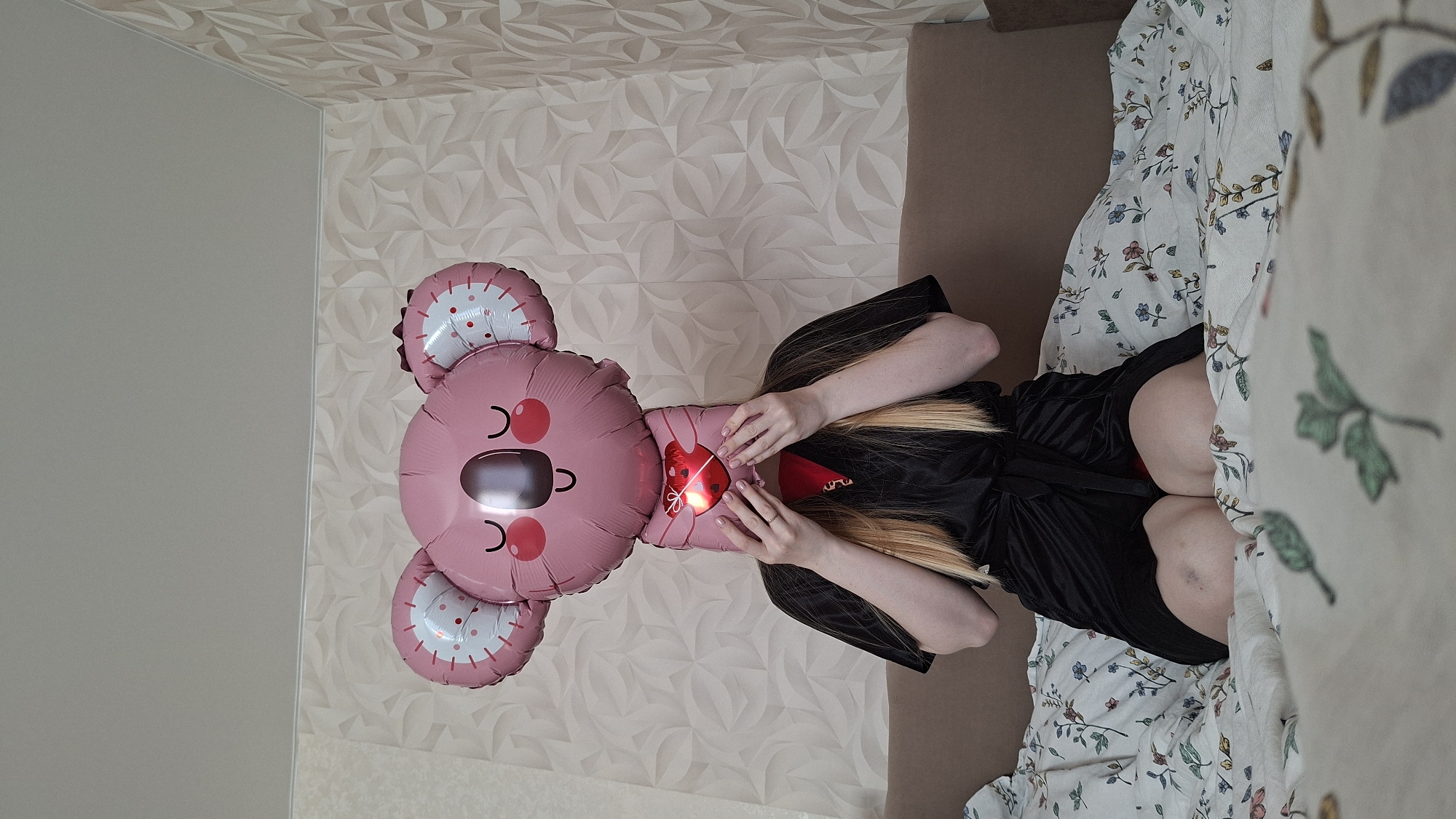 The image depicts a woman in a black robe lying on a bed covered with floral-patterned bedding. She is holding a pink koala-shaped balloon with a red heart in the middle. The background consists of a wall with a textured, geometric pattern. The location is likely Chaikovsky, Russia.