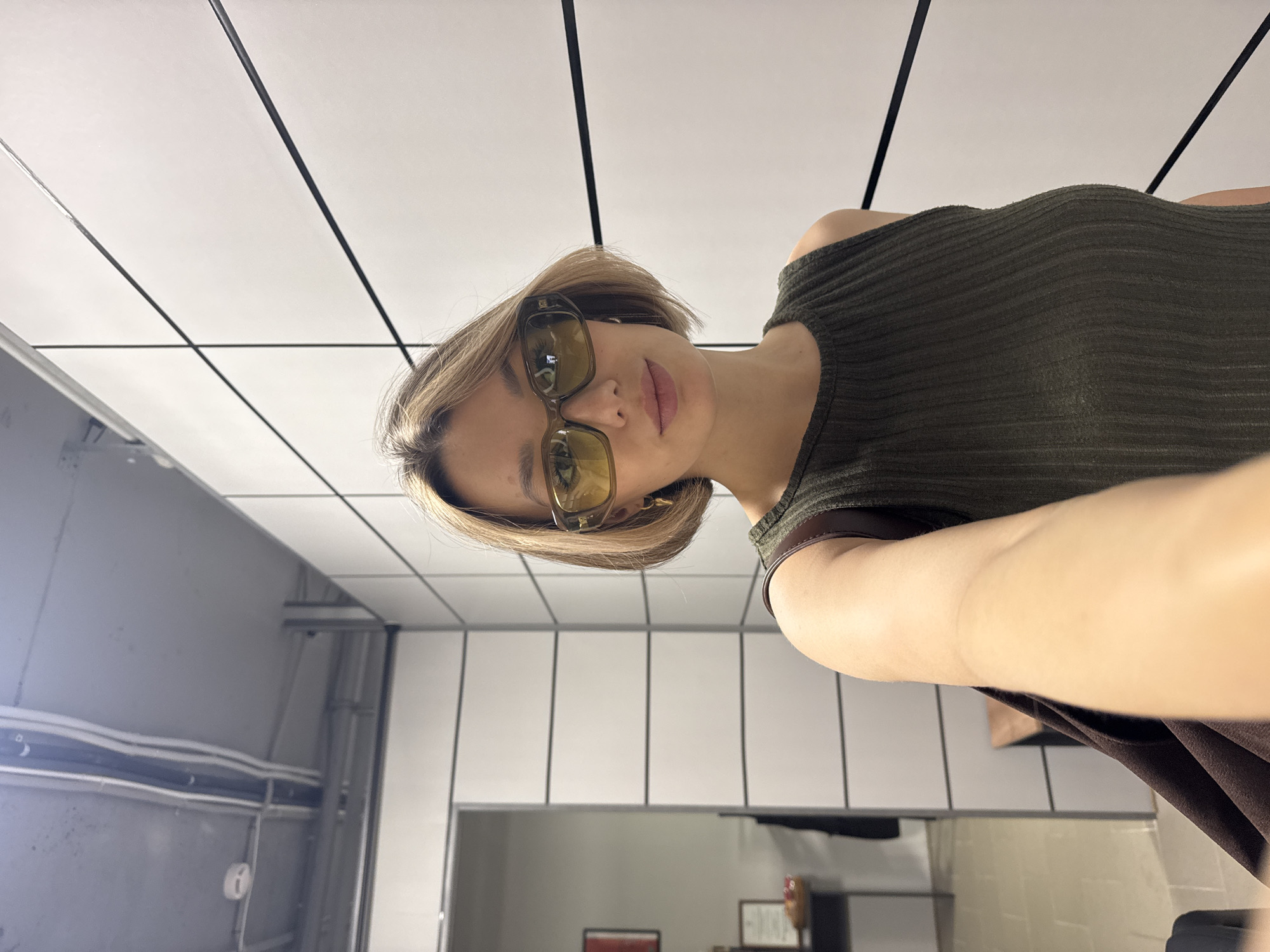 An over-the-shoulder selfie is taken by a young Caucasian woman with shoulder-length blonde hair, wearing large, square-framed olive-green sunglasses and a dark green ribbed sleeveless top. She is standing in what appears to be a fitting room or changing area, evidenced by the tiled walls with horizontal black lines and the doorway to another room visible to her left. The environment features exposed conduit and lighting on the ceiling, and a bench and what seems to be a display rack in the background. The woman's right arm is extended, holding the camera, and a dark brown bag is slung over her left shoulder. The overall lighting is bright and diffused.
