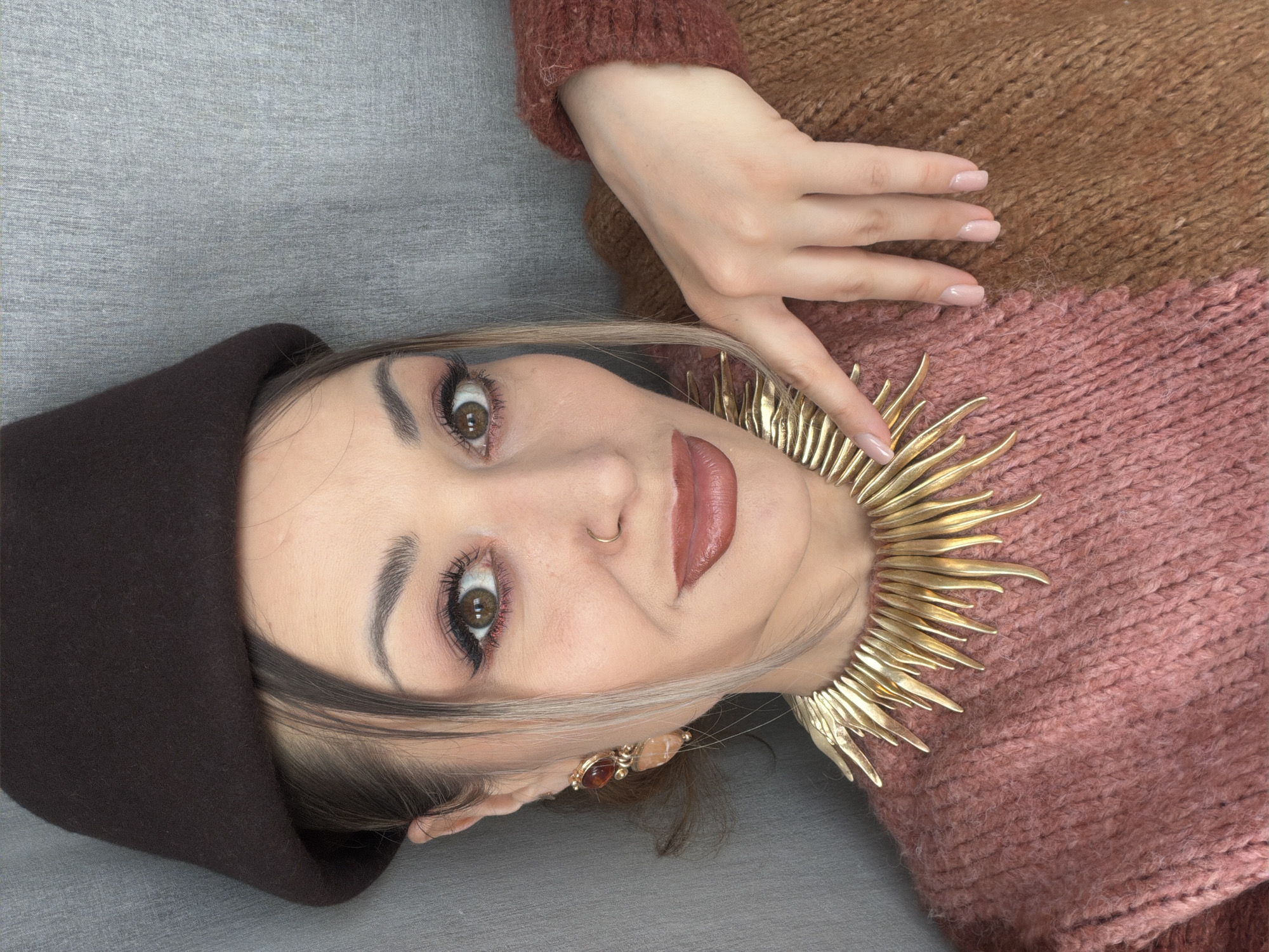 A woman is lying on a gray surface, wearing a brown hat and a color-blocked brown and pink sweater. She is also wearing a large gold necklace with a spike-like design. Her right hand is resting on her sweater. She has fair skin, brown eyes, and is wearing makeup. A small gold nose ring is visible. The image does not contain information to identify the location within Krasnoyarsk, Russia.