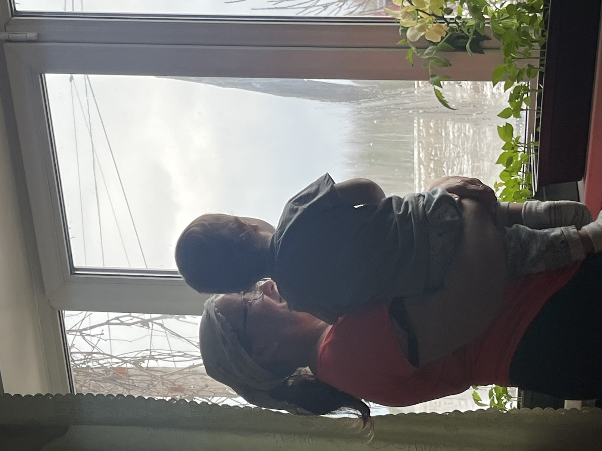 A woman with short gray hair, wearing a coral-colored shirt and black pants, holds a young child in front of a window. The child is facing away from the camera, wearing a gray shirt and gray pants. The window looks out onto a rainy day, obscuring the view of the outside, and there are plants on the windowsill. The location is Сосново, Russia.