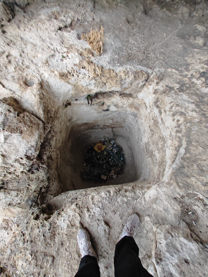 The image depicts a deeply cut, square-shaped rock well or cistern, likely an ancient or historical structure, within the vicinity of Shumen, Bulgaria, possibly on the Shumen Plateau. The pit is observed from a high-angle perspective, looking directly down into its depths.
The primary subject is a rock-hewn cavity with relatively smooth, light-colored vertical walls, approximately 2-3 meters deep. The base of this cavity is filled with a dense accumulation of refuse. This refuse consists predominantly of dark-colored glass bottles, intermingled with various plastic items and lighter-colored debris, including a visible crumpled yellow plastic bag.
The ground immediately surrounding the pit is composed of irregular, light grey-beige natural rock, showing signs of erosion and natural weathering. Scattered dry vegetation and sparse plant growth are visible in the rocky terrain beyond the immediate edge of the pit.
In the lower foreground of the image, the lower legs and feet of an individual are visible, positioned at the edge of the pit. The individual is wearing dark trousers and light grey or beige athletic shoes with white soles and laces. The footwear suggests a modern presence at an historical or natural site. No other individuals are visible. No actions or interactions beyond observation are depicted.