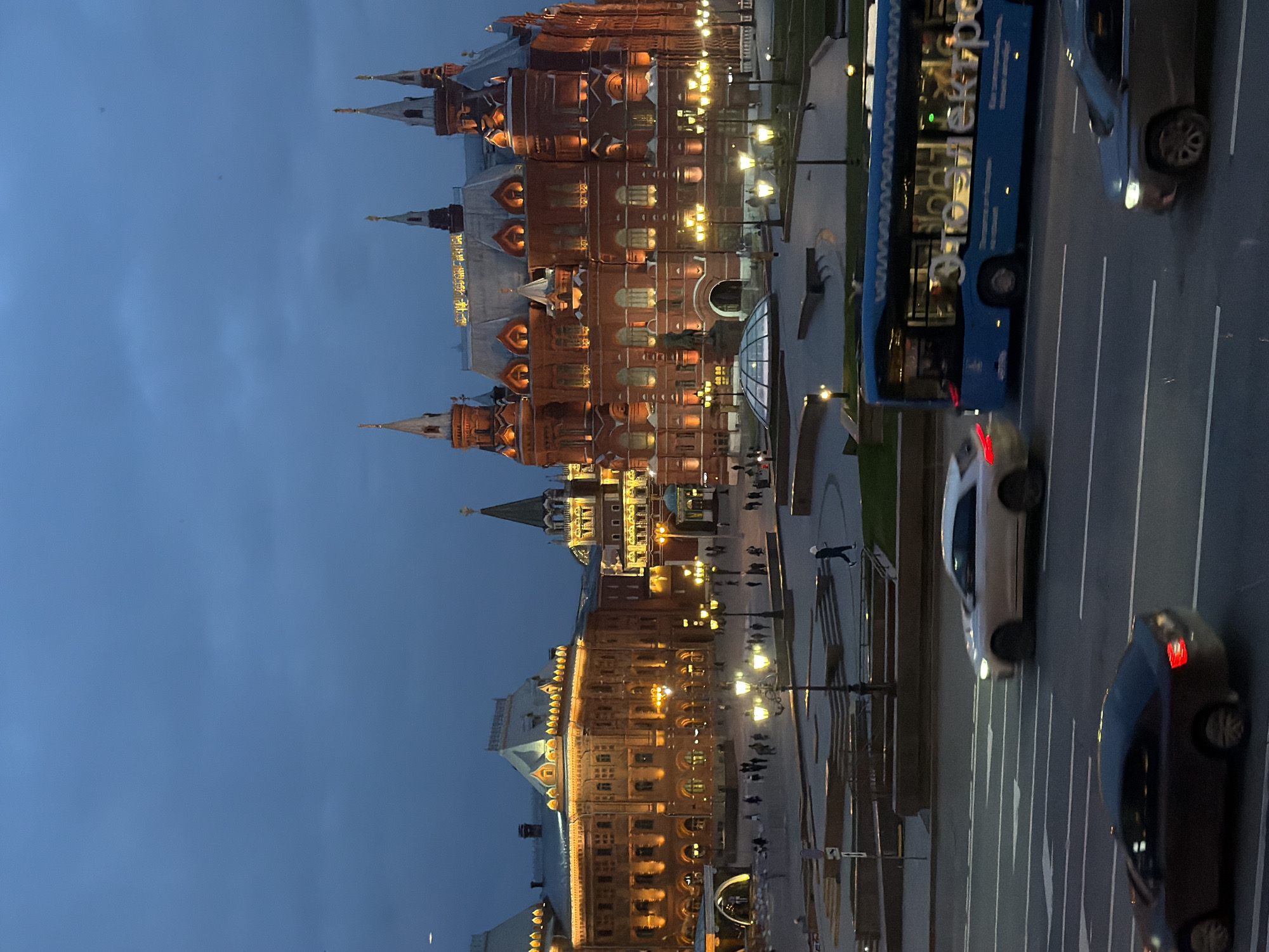 The image shows a night scene in Moscow, Russia, featuring the illuminated State Historical Museum and other buildings along Red Square. Several cars and a bus are visible on the road adjacent to the square. Pedestrians are walking along the sidewalk. The sky is a deep blue, indicating twilight or early evening.