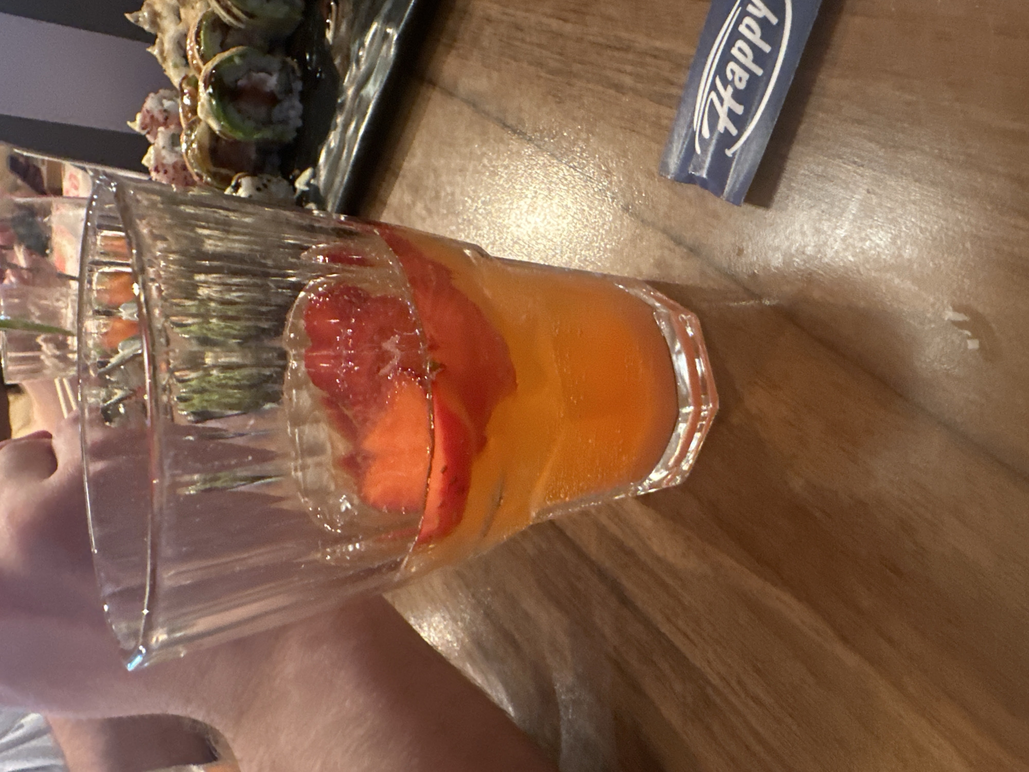 A close-up photograph captures a clear, ridged glass filled with an orange beverage and several sliced strawberries, positioned on a light brown wooden table. The drink contains small bubbles, indicating carbonation. To the immediate right and slightly below the glass, a small blue packet with the word "Happy" in white script is visible. In the upper left background, the bare arm and shoulder of a person wearing a white garment are partially visible. To the right background, a black rectangular plate holds multiple pieces of sushi, some topped with a dark sauce and others with red or white sprinkles. Another partially obscured glass is present further in the background. The wooden table exhibits a visible grain and reflections from an overhead light source. The scene depicts a dining environment in the city of Unknown, Unknown.