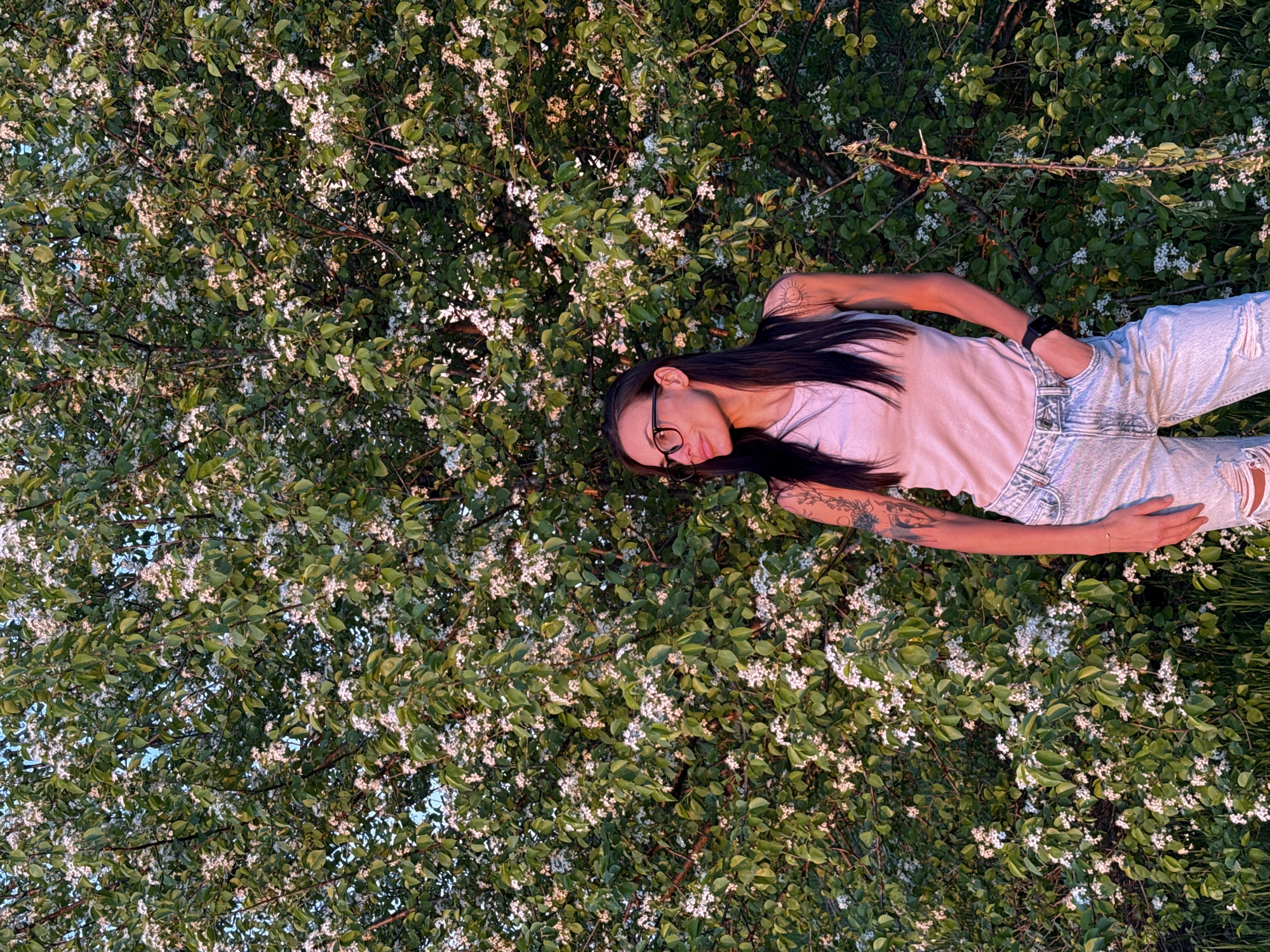 A young woman lies amongst flowering foliage in what appears to be Pleven, Bulgaria, based on the request. She wears jeans and a pink t-shirt and glasses. The flowering foliage is lush and green with small white flowers. Her arms are resting on the foliage near her body. She has tattoos on her arms. She is wearing a watch.