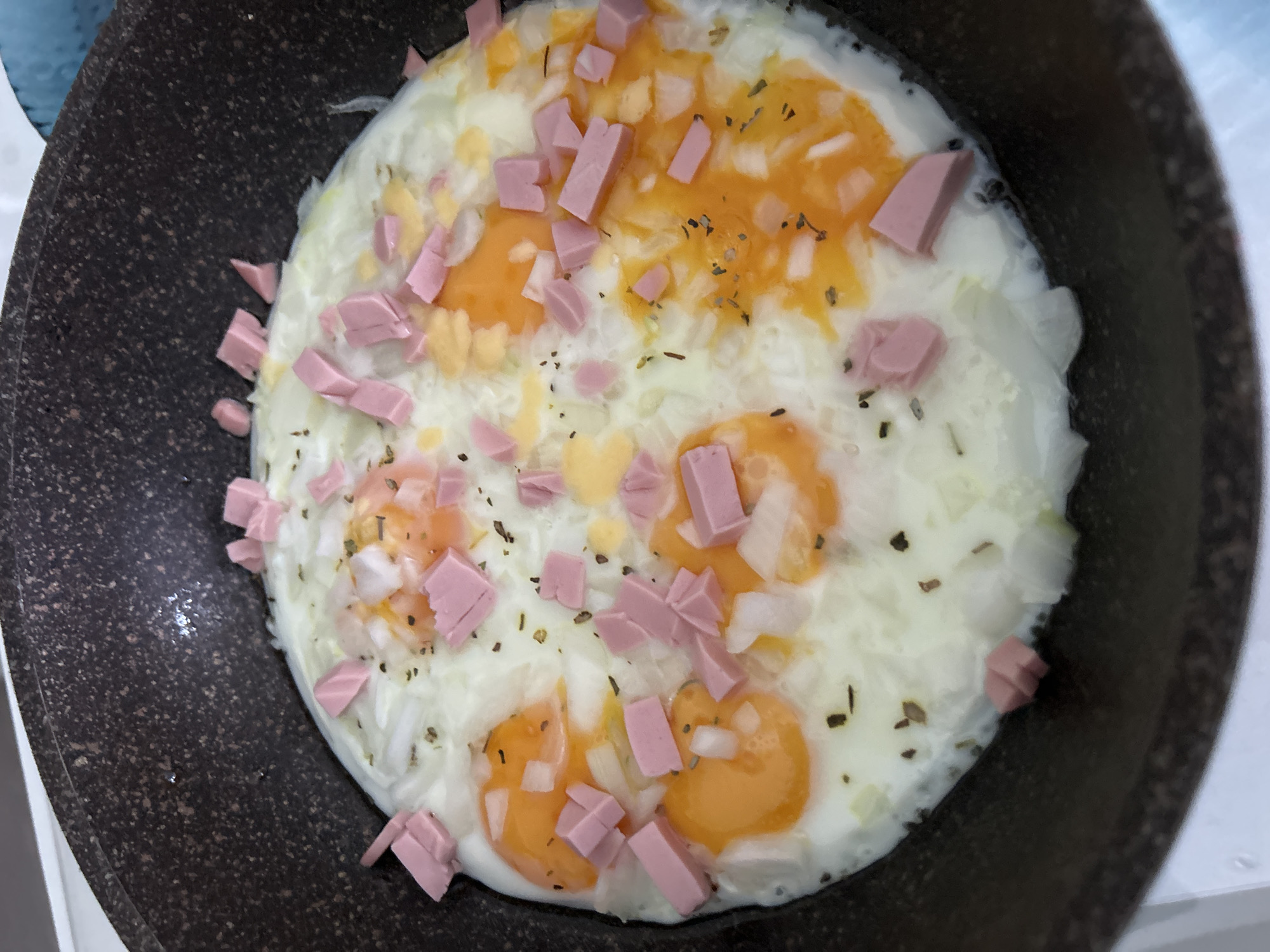 A close-up, top-down shot shows several fried eggs with visible yolks in a dark-colored frying pan. The eggs are topped with diced white onions, small pink cubes of ham or processed meat, and specks of dried herbs, possibly oregano or parsley, and black pepper. The frying pan has a speckled texture, typical of a non-stick coating. The background is blurred, but suggests a white surface, possibly a countertop or table. There is no indication of the city of Noviy, Russia, or any specific location within it.