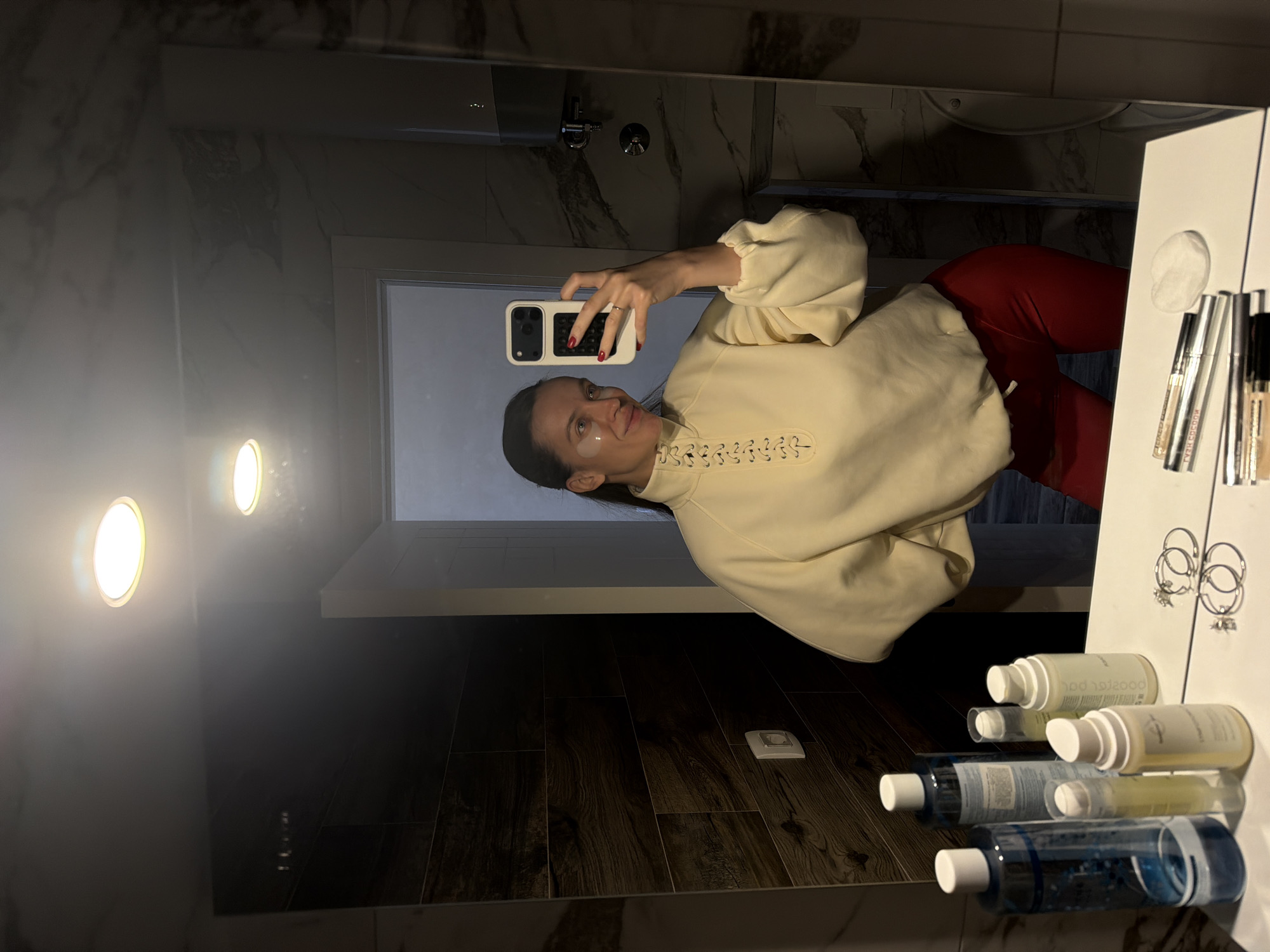 A person is taking a selfie in a bathroom with wood-paneled floors, white walls with marble accents, and vanity lighting. The person is wearing a cream-colored sweatshirt with lace-up detailing at the neck and red pants. A white vanity is visible with cosmetic products and jewelry. The bathroom is located in an interior space. The city of Budva and the country of Montenegro are not evident in this image.