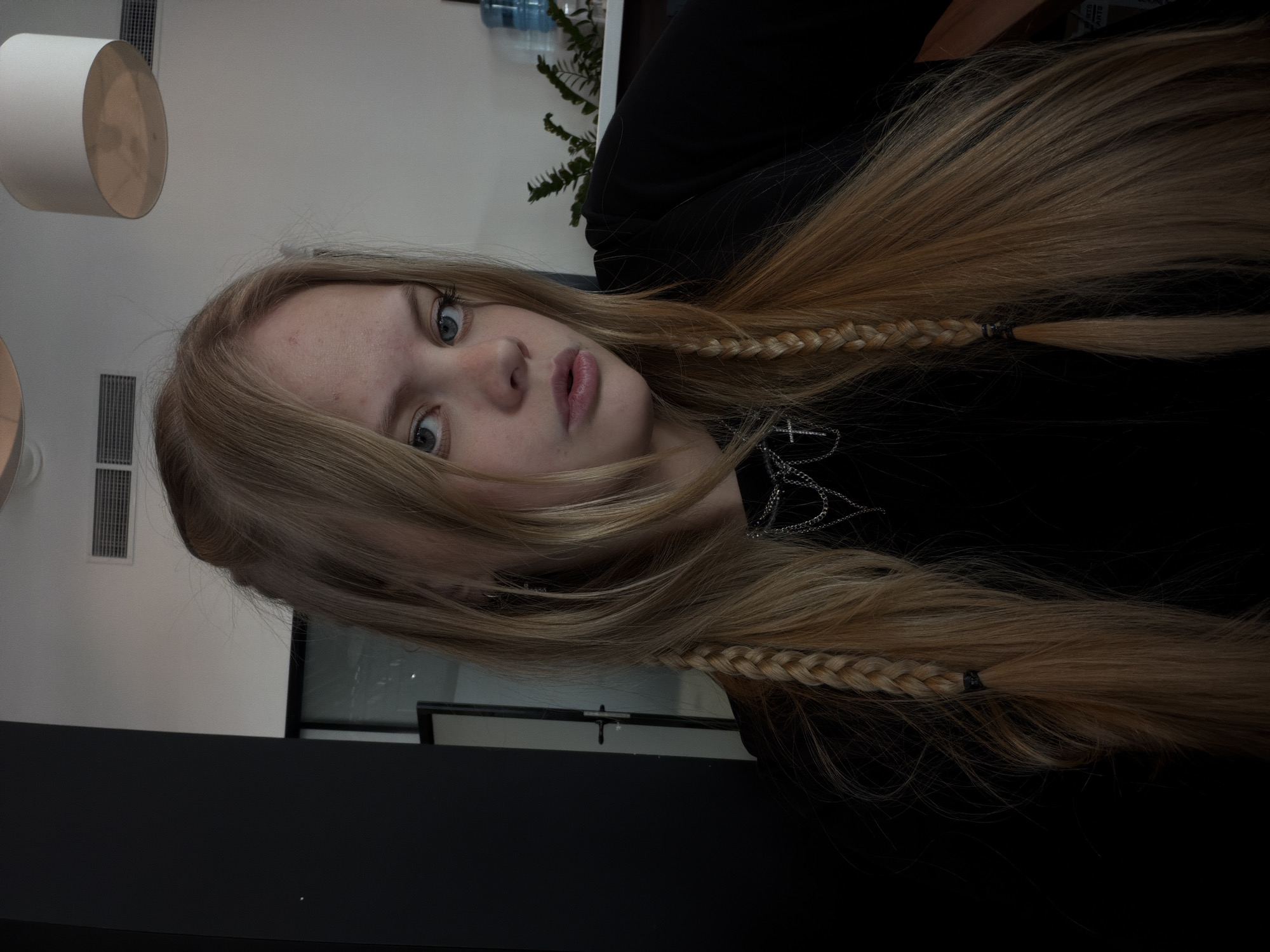 A head-and-shoulders portrait captured indoors features a young person with long, straight blonde hair and light blue or grey eyes, looking directly into the camera. The subject has fair skin and a neutral expression, with lips slightly parted. Their long hair is styled with two thin braids woven into the front sections, framing the face. They are wearing a dark, likely black, top, and a silver chain necklace with a cross pendant is visible on their chest.
The background suggests a modern interior space. A white ceiling with rectangular air vents and two cylindrical, off-white light fixtures is visible. Behind the subject, there is a dark green leafy plant and several plastic water bottles on a shelf or surface. A dark, flat surface, possibly a desk or table, occupies the foreground. The lighting appears to be indoor ambient light, contributing to a somewhat dim or underexposed appearance of the photograph. The person is posing for the camera, with no specific activity taking place. There is no visible text in the image.