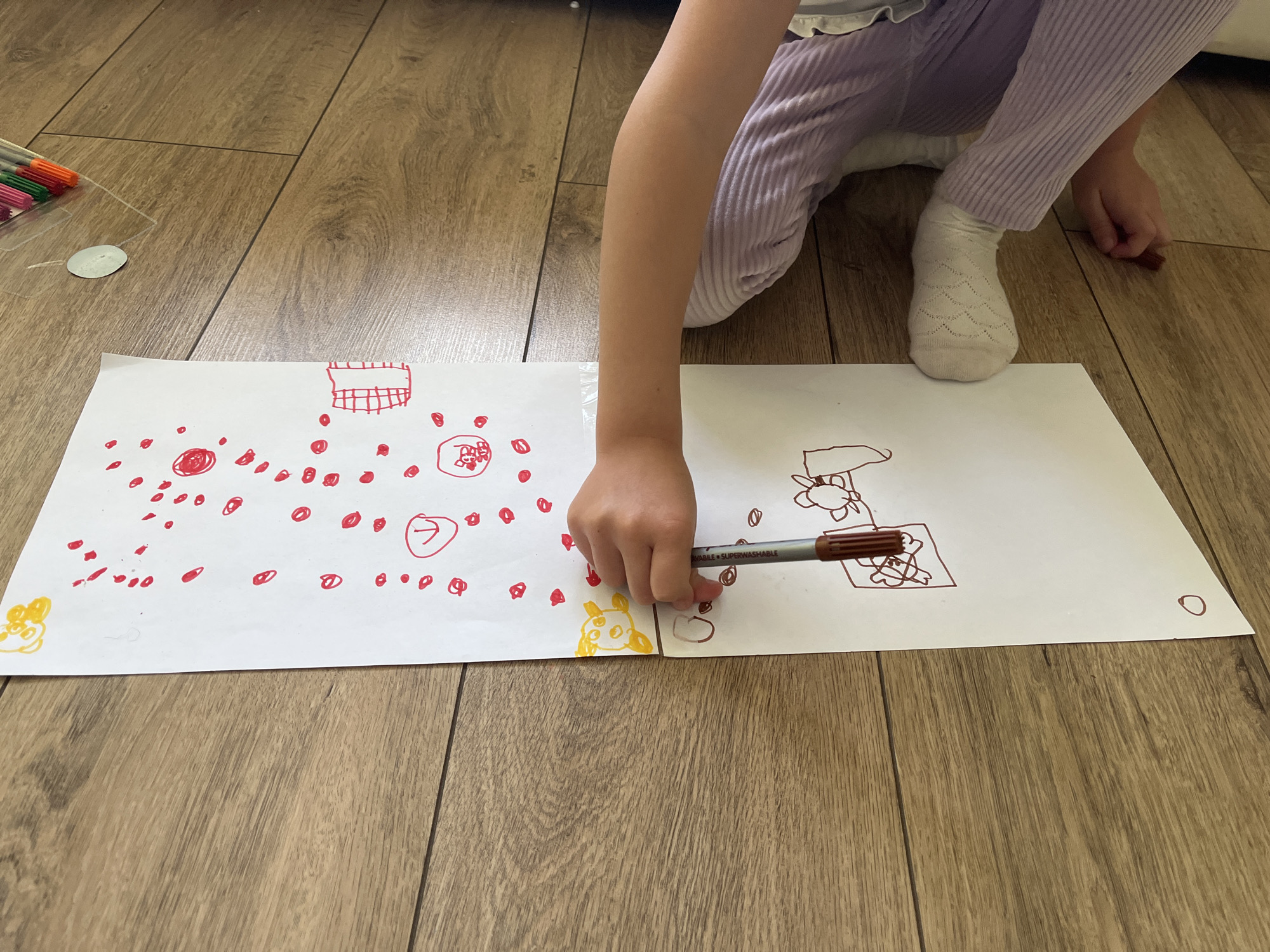 A child is drawing on two pieces of white paper that are laid out side-by-side on a wooden floor. The child is kneeling, wearing light purple striped pants and a white sock on one foot. The left side of the drawing features numerous red dots and a yellow drawing, possibly of a cartoon character. On the right side, the drawing is done in brown ink, depicting abstract figures. The child is holding a brown marker in their right hand, as if in the process of adding to the drawing on the right side. A set of colored markers is visible to the left, along with a small round object. The scene is indoors, with a focus on the child's activity. The location is Unknown, Unknown.