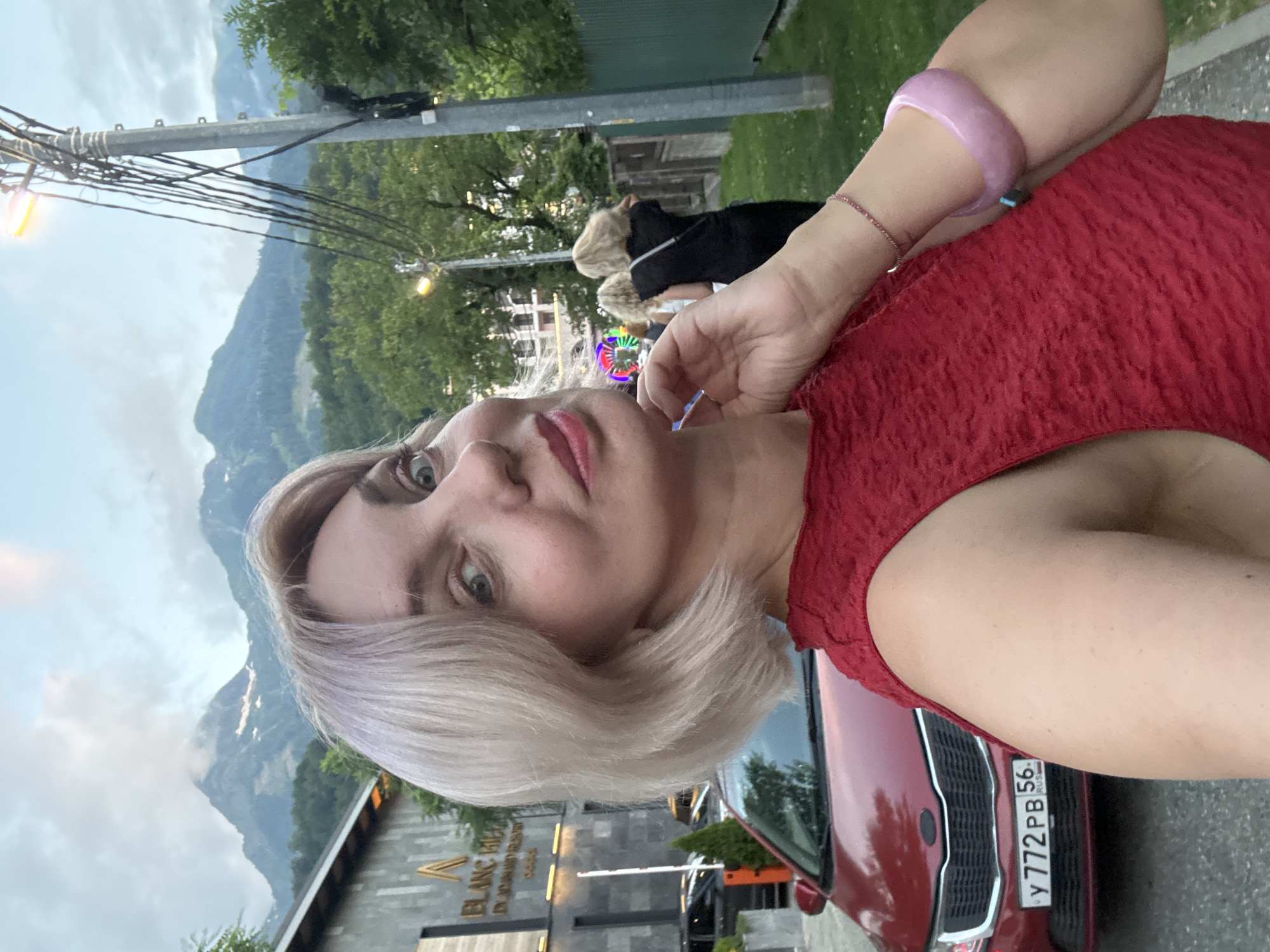 A selfie captures a fair-skinned Caucasian woman with blonde hair styled in a bob, wearing a red textured sleeveless dress. She has light blue eyes and is looking slightly to her left, with her right hand touching her neck. A pink beaded bracelet and a translucent pink bangle adorn her right wrist. The background features a modern, multi-story building with the text "BLANC RIED" visible on its facade. To the left, a red Kia Rio car is parked, displaying a Russian license plate that reads "Y 772 PB 56". Beyond the car and building, lush green trees and mountainous terrain under a cloudy sky are visible, suggesting an outdoor setting in a mountainous region. In the mid-ground, two women with blonde hair are walking away from the camera, one wearing a black dress. A streetlamp with illuminated lights and a tangle of wires stands to the right of the woman. The scene appears to be in the city of Estosadok, Russia, a resort town known for its mountainous setting.