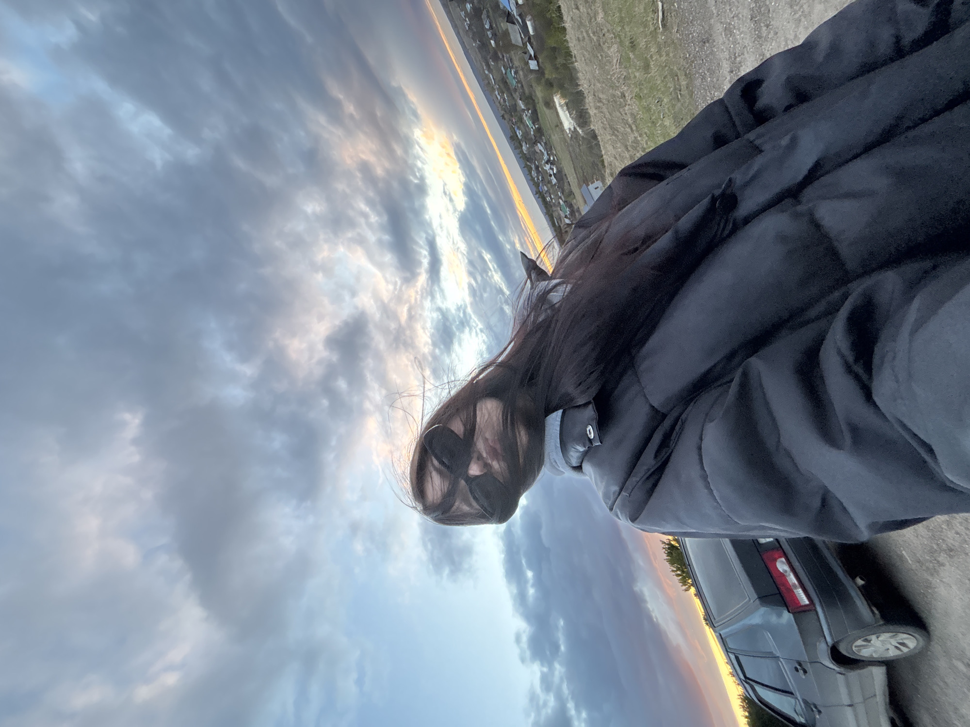 A photograph, presented on its side, captures an outdoor scene at twilight, likely in Фоки, Russia. The main subject is a person, likely a woman, positioned in the foreground. Her dark, long hair cascades over her face, obscuring most features, and she appears to be wearing dark sunglasses. She is dressed in a dark, possibly black or navy, puffer jacket over a lighter grey layer.
The expansive background is dominated by a dramatic sky filled with scattered clouds, illuminated by the warm glow of a setting or rising sun, displaying vibrant orange, yellow, and blue hues at the horizon. Below the sky, in the midground, a small village or settlement with numerous houses and some vegetation is visible along a distant landmass or shoreline.
To the lower right, the rear portion of a dark-colored car, showing a taillight and part of a wheel, is parked on what appears to be a dirt or gravel path. The overall atmosphere suggests a quiet, expansive landscape during the transition from day to night or night to day. No visible text is present.
