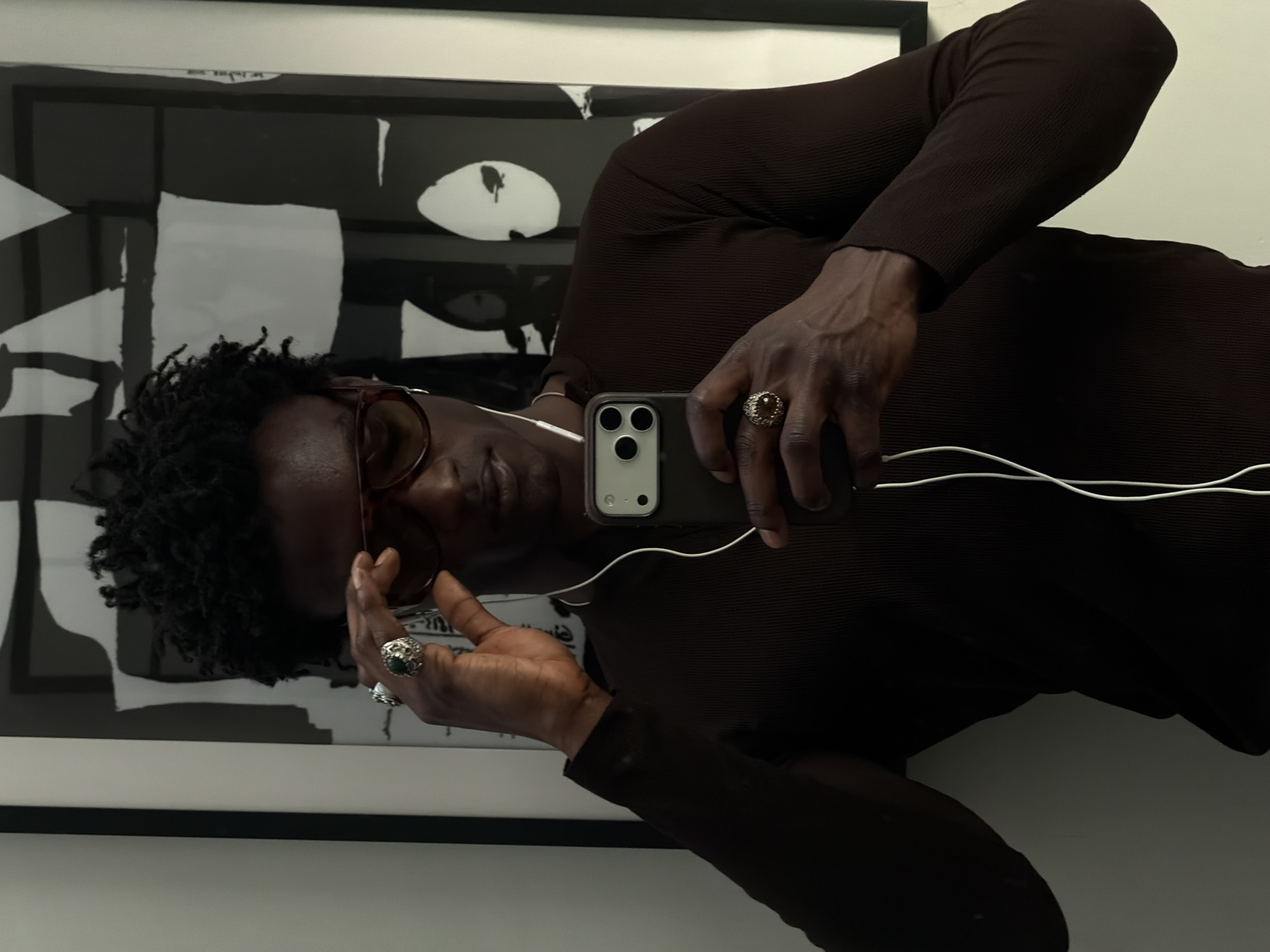 A dark-skinned person is taking a selfie, holding a smartphone with a triple-lens camera. They are wearing glasses, headphones, and a brown long-sleeved shirt. The person is wearing two ornate rings on their fingers. Behind them is a framed black and white abstract artwork. The location is likely an indoor setting in London, United Kingdom, but specific details are not visible.