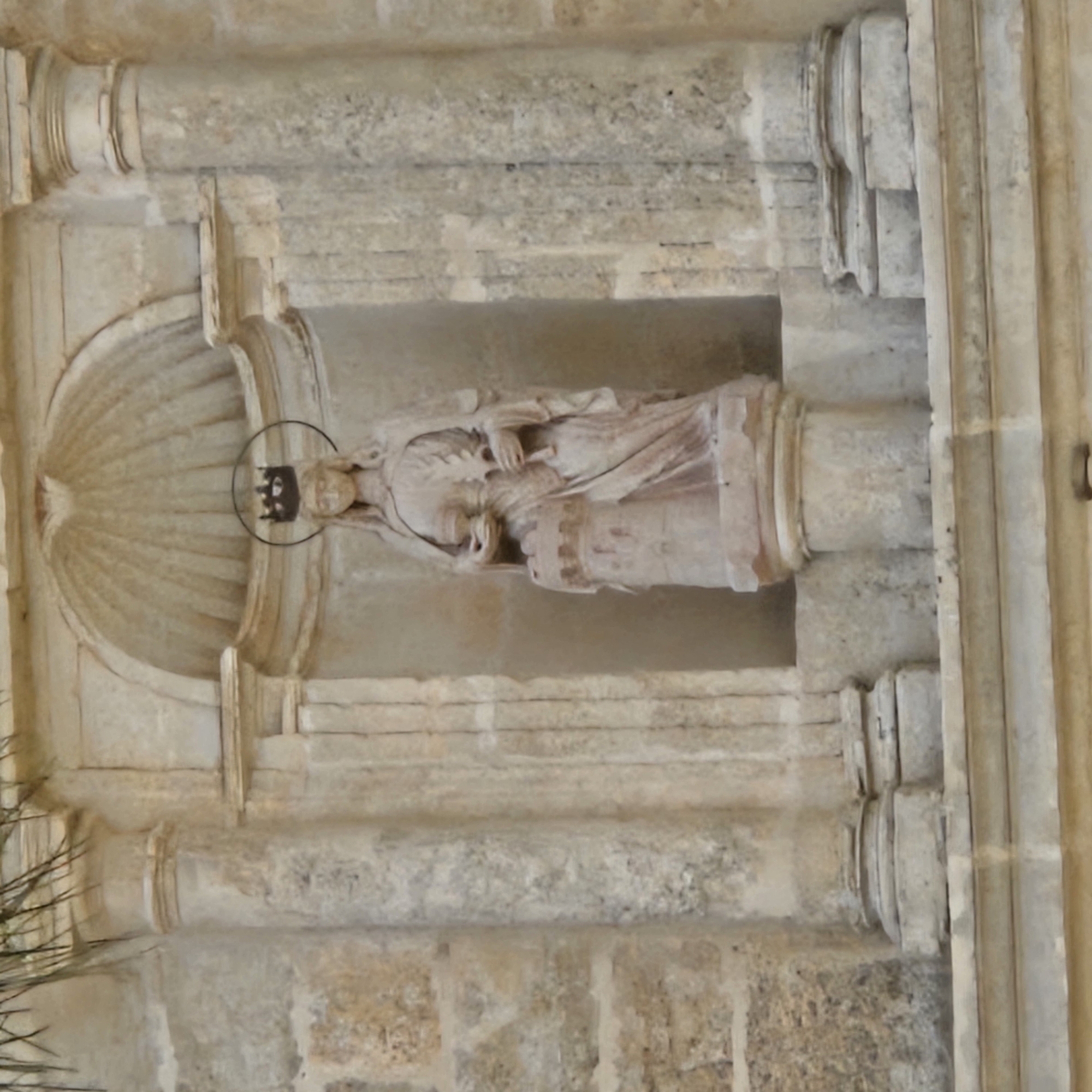 A stone sculpture of a figure lies horizontally within a decorative niche in a stone wall. The sculpture appears to be a woman holding an object; she wears a headpiece with a halo-like ring above. The niche has a semi-circular top section with radiating lines suggesting a clamshell shape. The surrounding wall is made of rough-hewn stone blocks and features architectural details such as columns and horizontal bands. The exact location in Algora, Spain is not discernible from the image.