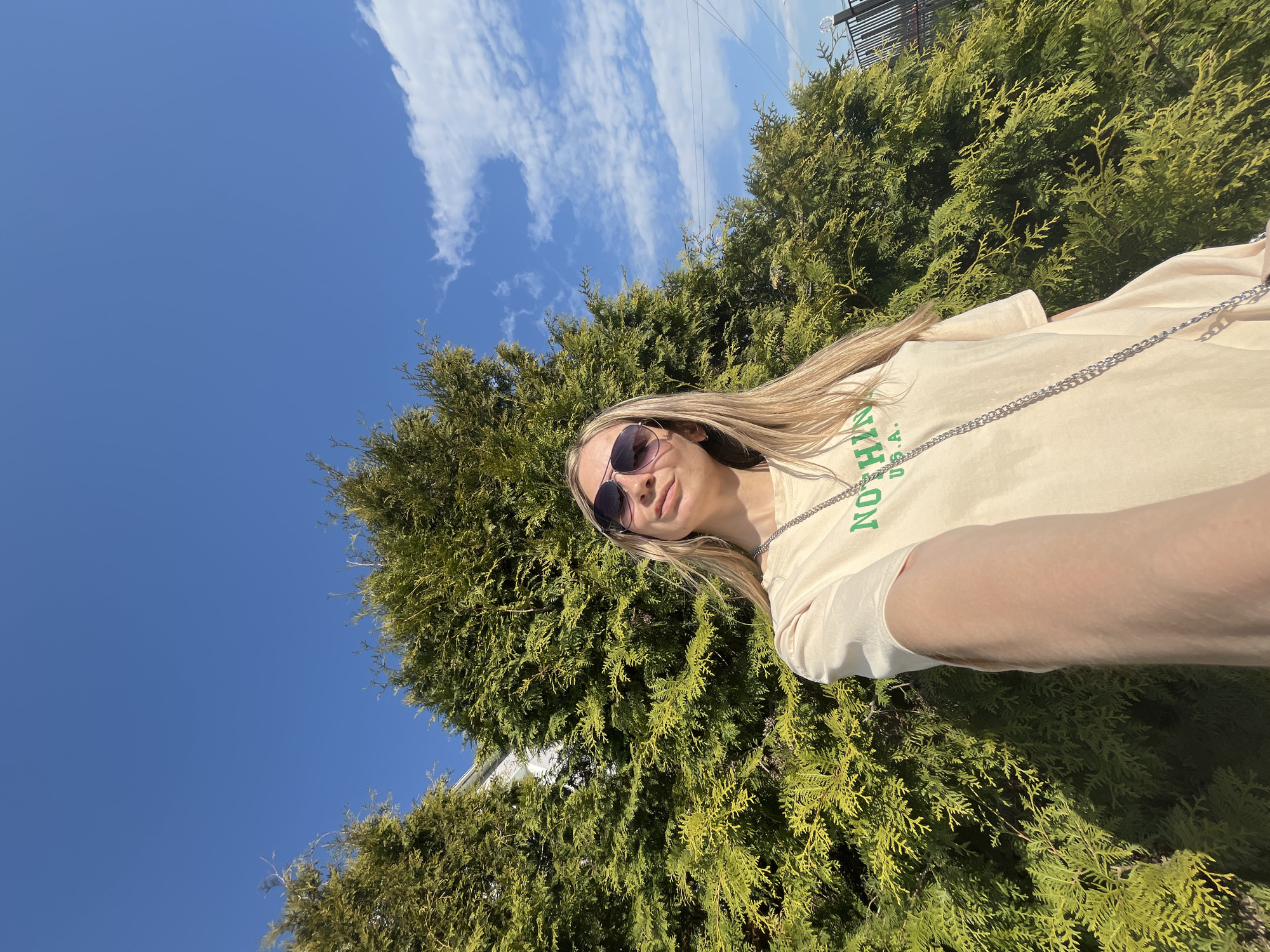 A woman with long blonde hair is posing for a selfie outdoors, with a backdrop of green trees and a bright blue sky with scattered clouds. She is wearing sunglasses and a light-colored t-shirt with green lettering. The perspective is from a low angle, looking up at the woman and the sky. The location is in Chaikovsky, Russia.