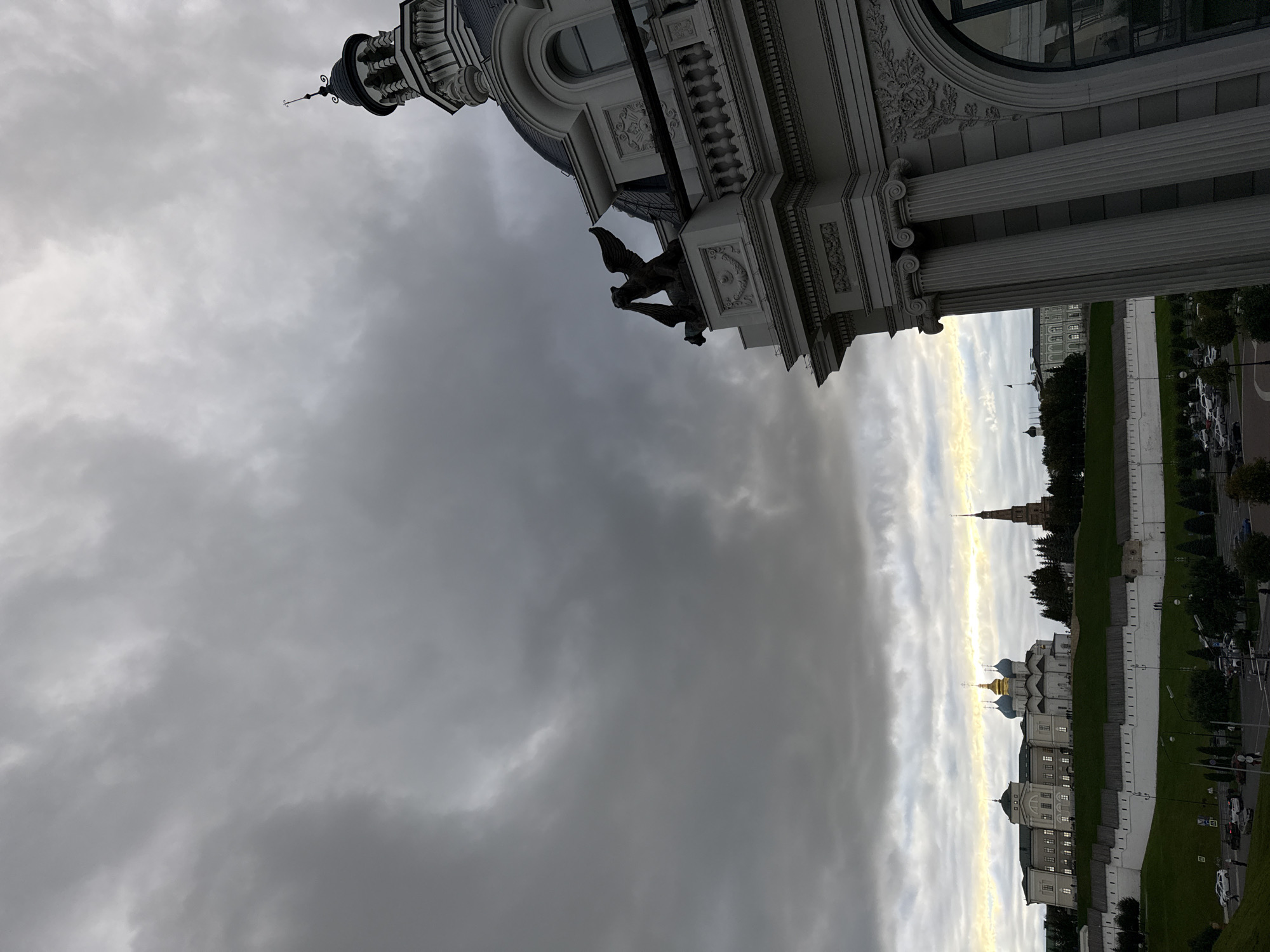 The image captures a view of the Kazan Kremlin from the National Gallery "Khazine" in Kazan, Russia, under a dramatic, cloudy sky. In the foreground, the ornate architecture of the National Gallery is visible, featuring classical columns and a domed tower. To the left, a white fortress wall, characteristic of the Kremlin, forms a prominent barrier along a grassy slope. Beyond the wall, the Kul Sharif Mosque, with its distinctive blue domes, and other buildings within the Kremlin complex, are visible. A tall, slender minaret stands to the right of the mosque. The sky is filled with dark, heavy clouds, suggesting overcast weather, with a sliver of yellow light breaking through on the horizon. The scene appears to be a street-level view with cars and pedestrian activity hinted at in the lower portion of the image.