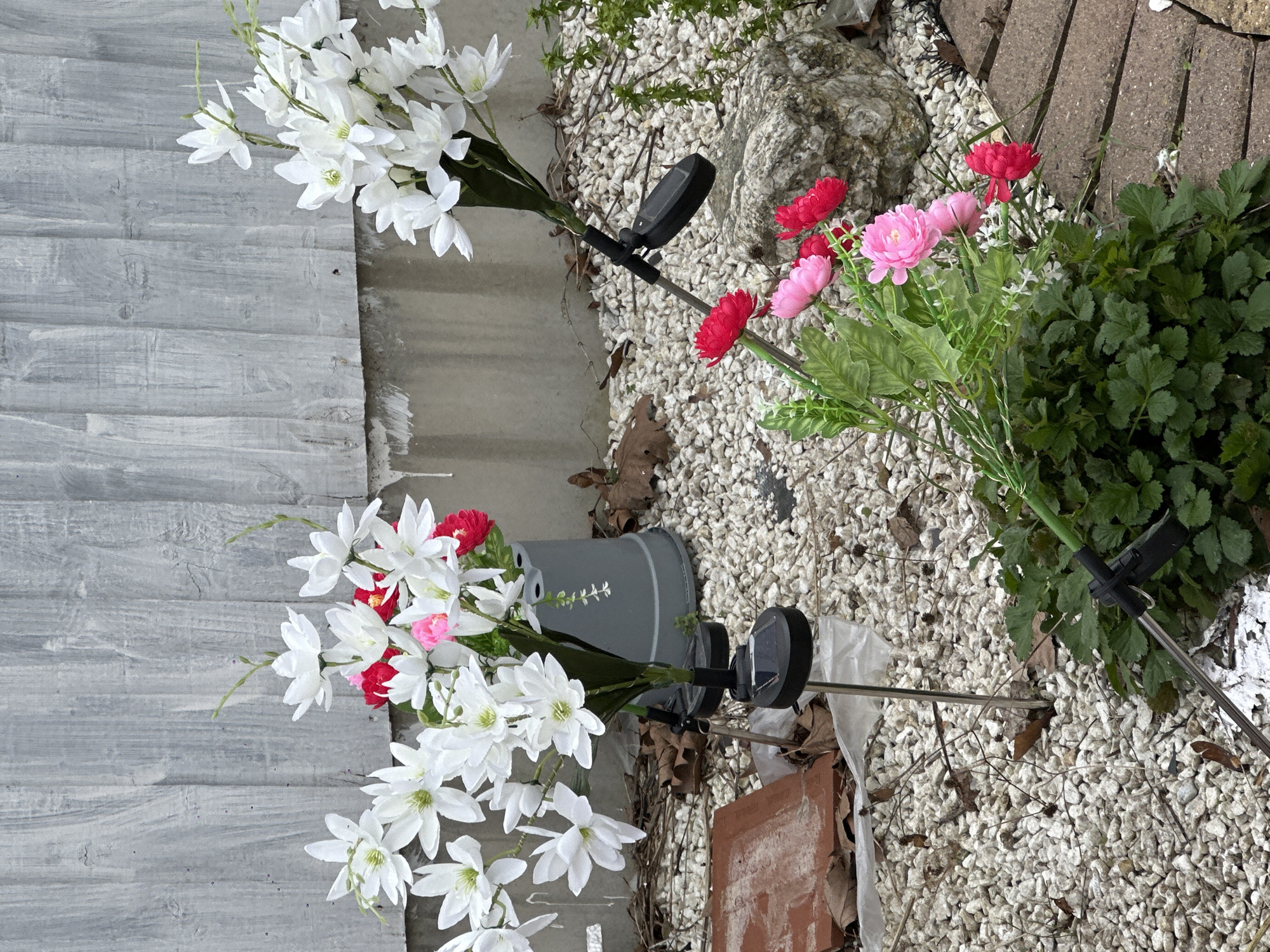 The image shows an outdoor scene with artificial flowers placed in a bed of white gravel. In the left foreground, a bouquet of white flowers with a few red and pink blooms is positioned near a gray-painted fence with a wood-grain effect. The flowers are affixed to metal stakes with black solar lights attached. Behind these flowers, a gray plastic pot is visible.
To the right of the white flowers, another set of artificial flowers in pink and red hues is arranged among green leafy foliage. These also have a similar stake and solar light setup.
The background features a mix of elements including a large rock, more gravel, and parts of a brick pathway. The scene is generally untidy with scattered leaves and debris. The presence of the fence and pathway suggests the location might be in a garden or similar residential outdoor space.
The image does not show people and provides no specific information that would allow determination of its location in London, United Kingdom.