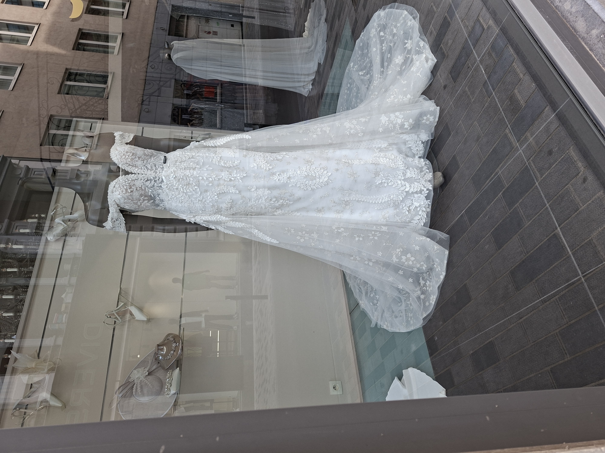 A white wedding dress is displayed on a mannequin in a storefront window, possibly in Brussels, Belgium. The dress is heavily embellished with lace and has a long train. The reflection in the window shows a building with multiple floors and windows, as well as a street scene with other shops and passersby. Inside the store, other dresses and accessories, including shoes and a hat, are visible. The floor of the storefront appears to be tiled. The overall impression is a bridal shop showcasing its collection.