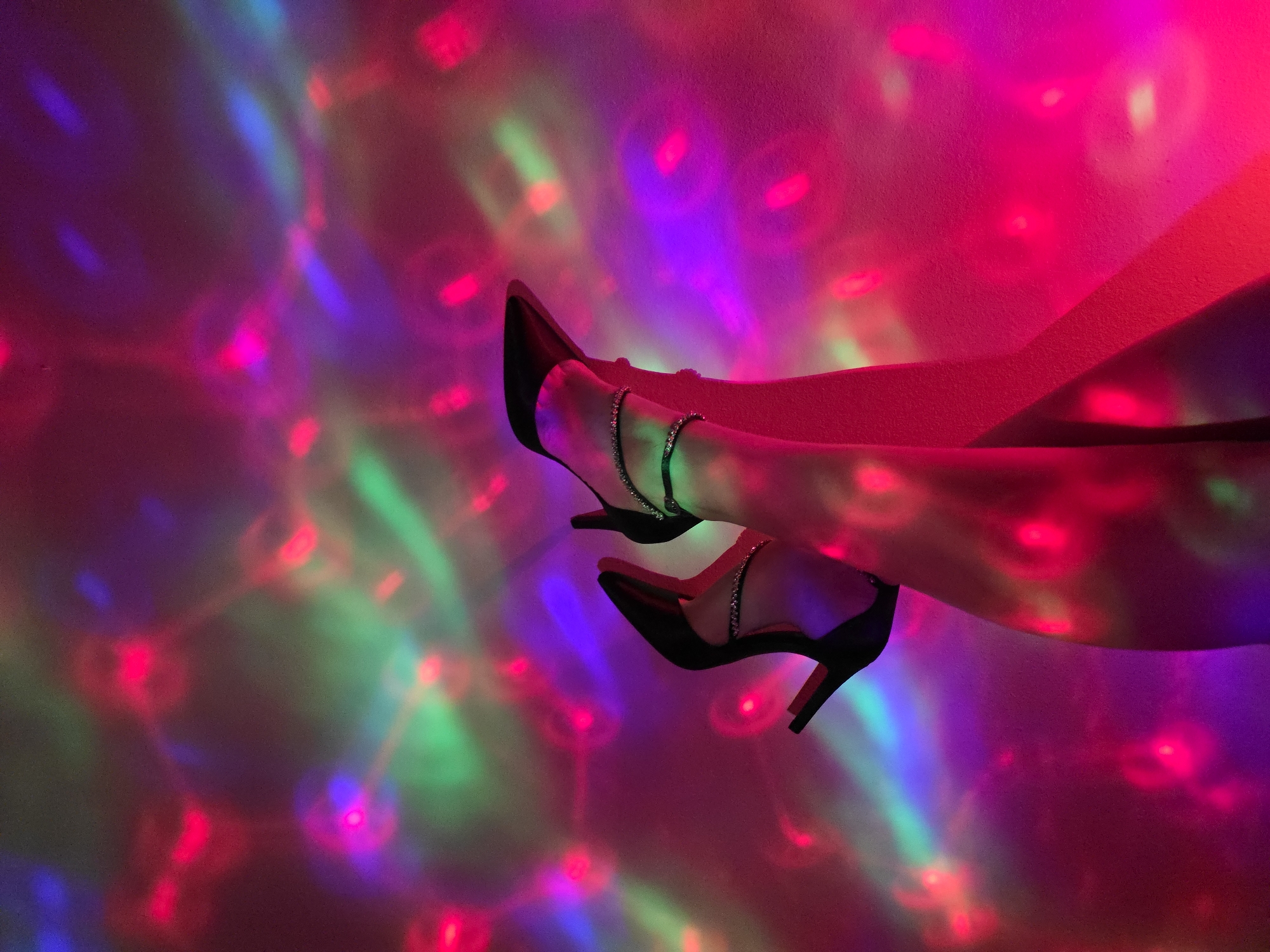The image depicts a pair of crossed legs, adorned with black high heels and anklets, set against a backdrop of colorful, blurred light patterns. The lighting is primarily red, pink, purple, green, and blue, creating a disco-like atmosphere. The location is unknown, as it is not clear from the image.
