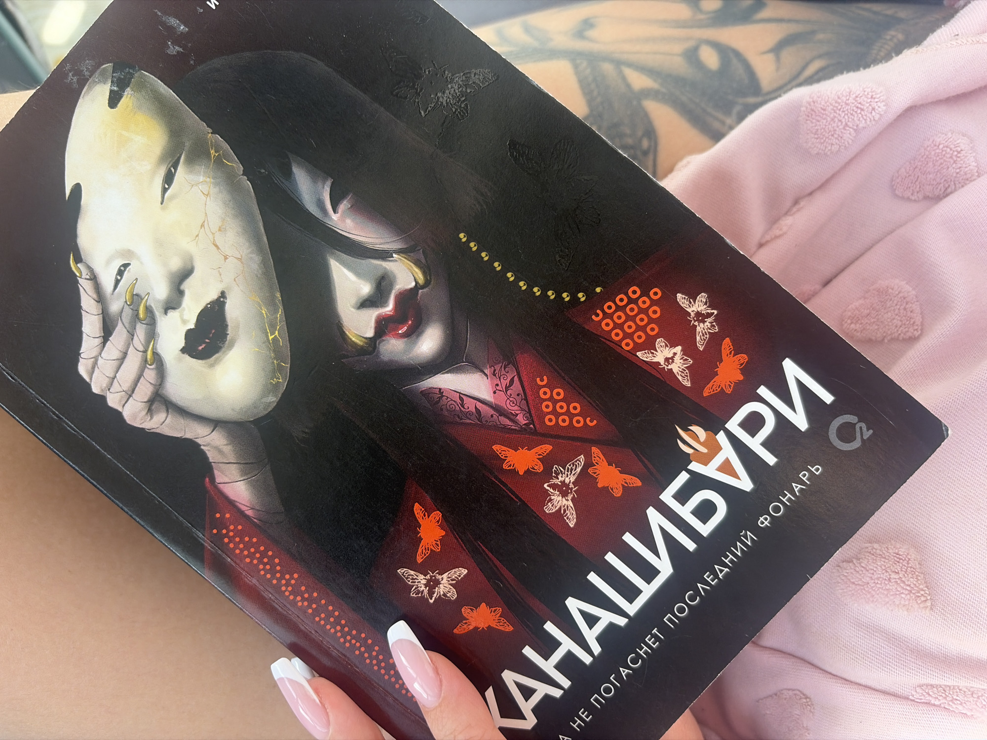 A book titled "Канашибури" (Kanashiburi) is held in a person's hand with French-tipped nails. The cover depicts a pale woman with dark hair, red lips, and gold embellishments, holding a cracked white mask with gold accents. The woman is dressed in a red and black kimono-style garment adorned with moth designs. The background is dark, with subtle, embossed moth patterns. The book's title and author information are printed in white. The book is resting on a light pink fabric with raised polka dot patterns. Part of a tattooed arm is visible in the background. There is no location information about Saint Petersburg, Russia.