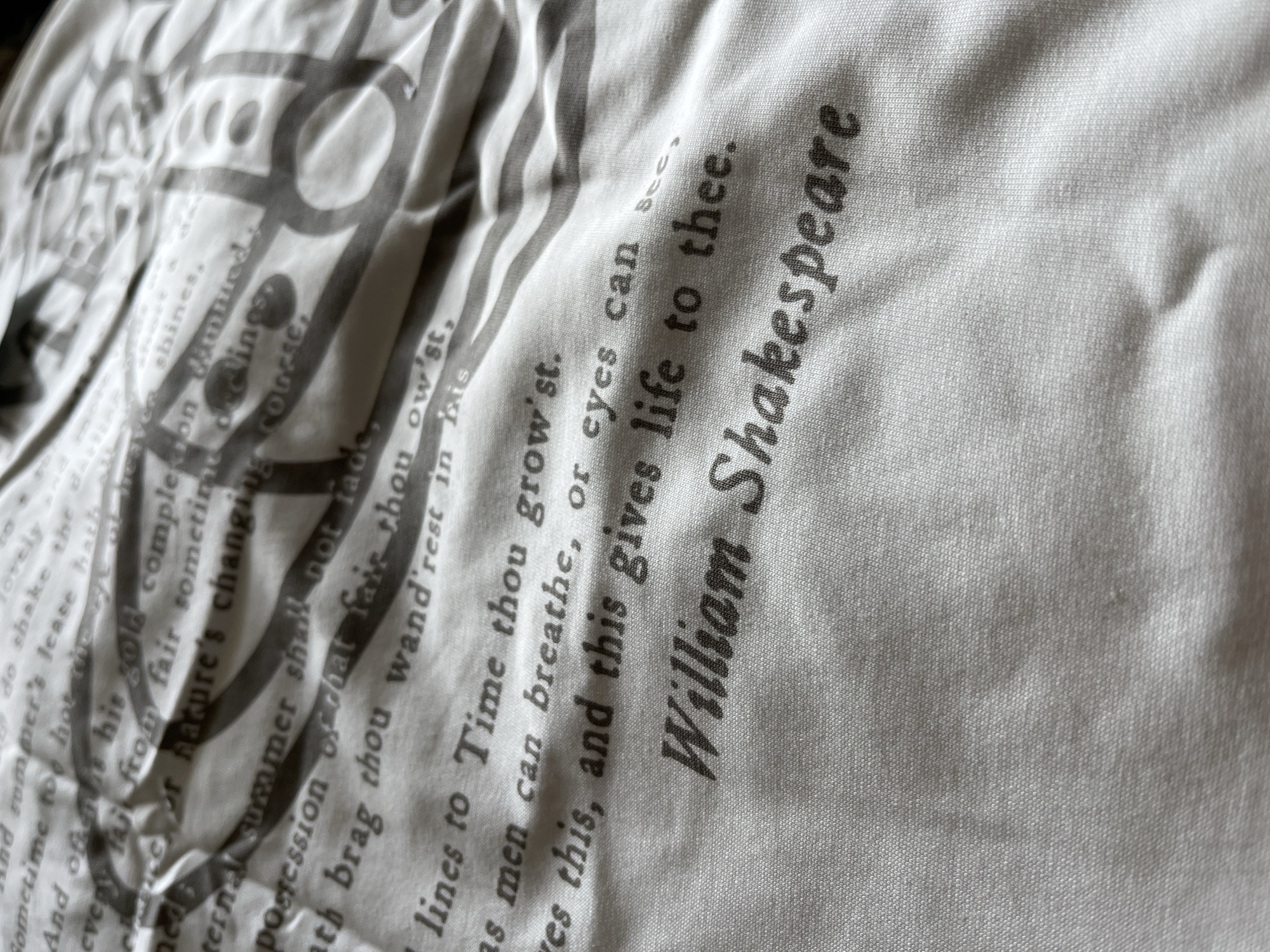 The image shows a close-up of a white fabric printed with text and graphic elements in shades of grey. The text includes phrases from Shakespearean sonnets, notably the line "So long as men can breathe, or eyes can see, So long lives this, and this gives life to thee," followed by the name "William Shakespeare." Other partial lines from sonnets are also visible. Interspersed with the text are swirling graphic design elements, creating a patterned effect on the fabric. The fabric appears to be wrinkled or folded, adding texture to the image. There is no indication that the image was taken in the city of Zlatograd, Bulgaria.