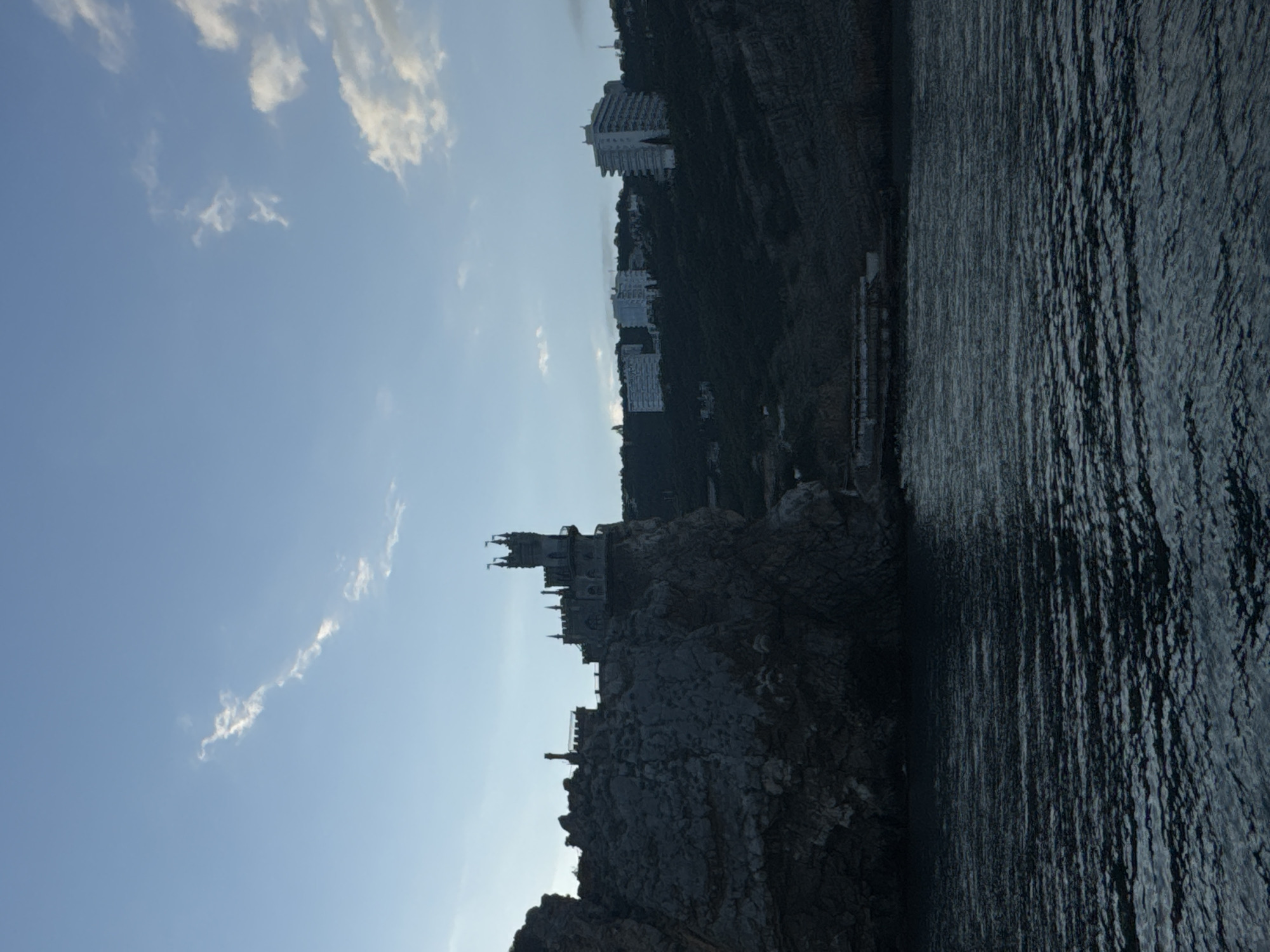 The image shows Swallow's Nest, a decorative castle located in Гаспринский поселковый совет, Ukraine. The castle is situated on a rocky cliff overlooking the sea. Behind the castle, further up the hillside, are several large buildings. The sky is blue with some clouds. The sea is dark and appears to be calm. The image is oriented with the castle and sea on the right side.