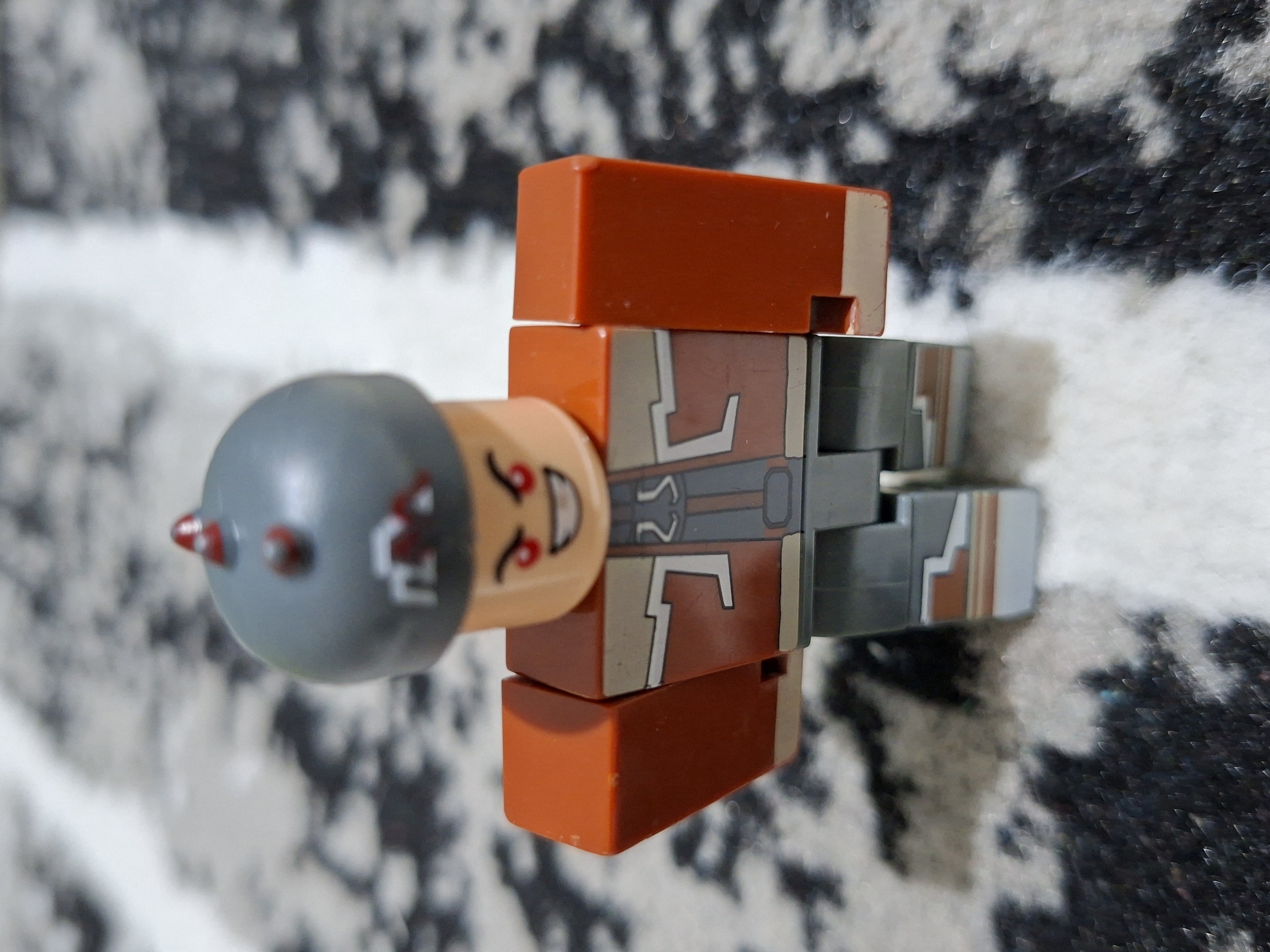 The image shows a LEGO minifigure lying on a carpet with a mottled white and black pattern. The minifigure has a grey helmet with red spikes, an orange torso with white and grey markings, and grey legs with similar markings. The location is Kutaglumpang, Indonesia.