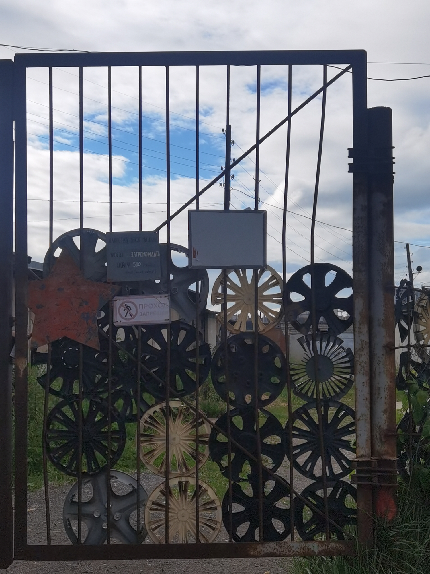 The image shows a metal gate decorated with car wheel rims and a red star. There are signs on the gate. One sign, in Russian, reads "WARNING. THIS TERRITORY IS PROTECTED. TRESPASSERS WILL BE PUNISHED. FINE 500 RUBLES." Another sign depicts a person being crossed out, indicating "NO ENTRY." The gate is located in the city of Chaikovsky, Russia, likely in an industrial or abandoned area, as suggested by the presence of electrical wires overhead and overgrown vegetation. The sky is cloudy.