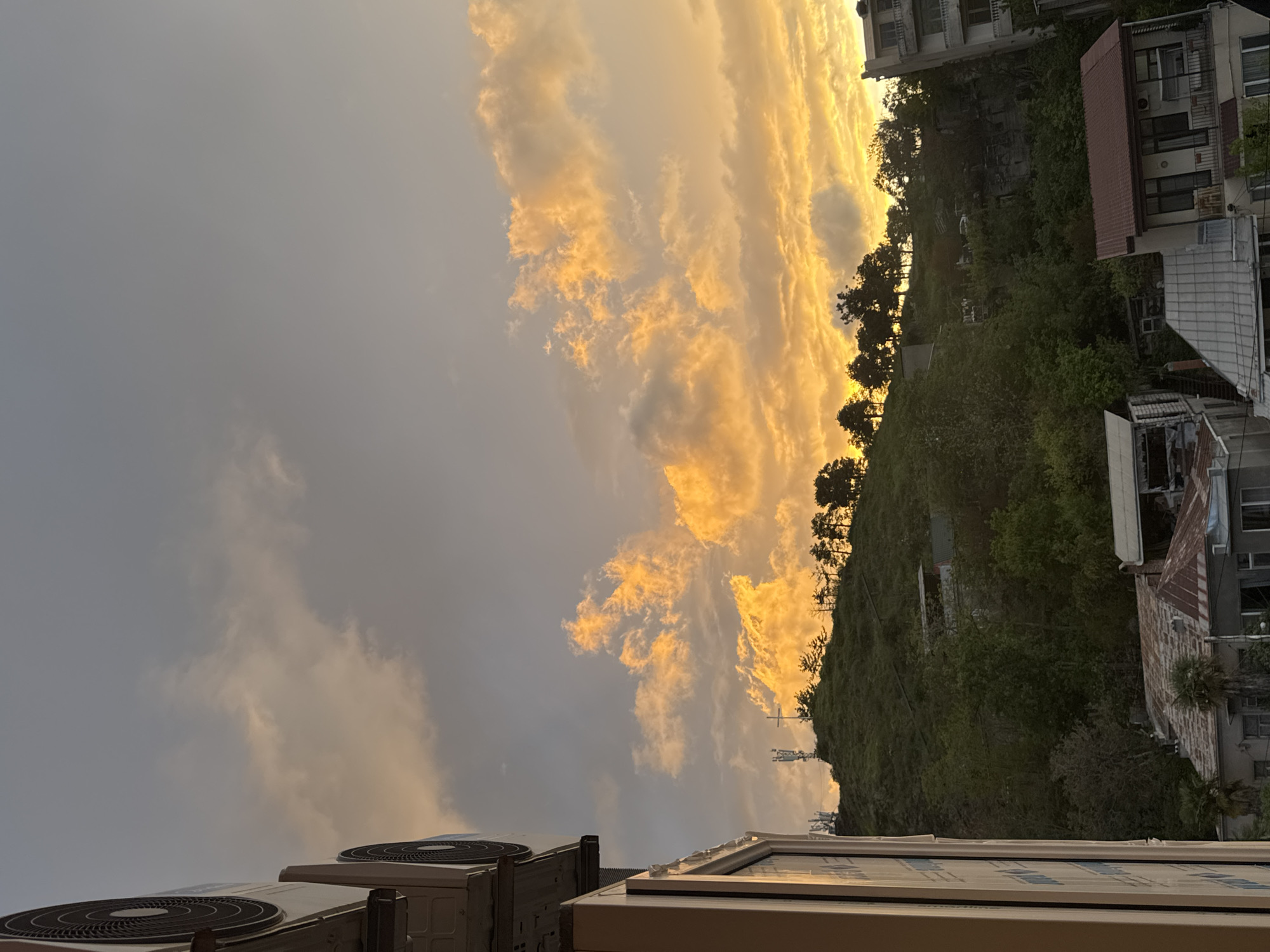 The image captures a sunset sky with orange-tinted clouds over a landscape featuring buildings, trees, and a hill. In the foreground, air conditioning units are visible. The scene appears to be taken from a high vantage point, possibly a rooftop or upper floor of a building in an urban area. The location is unknown.