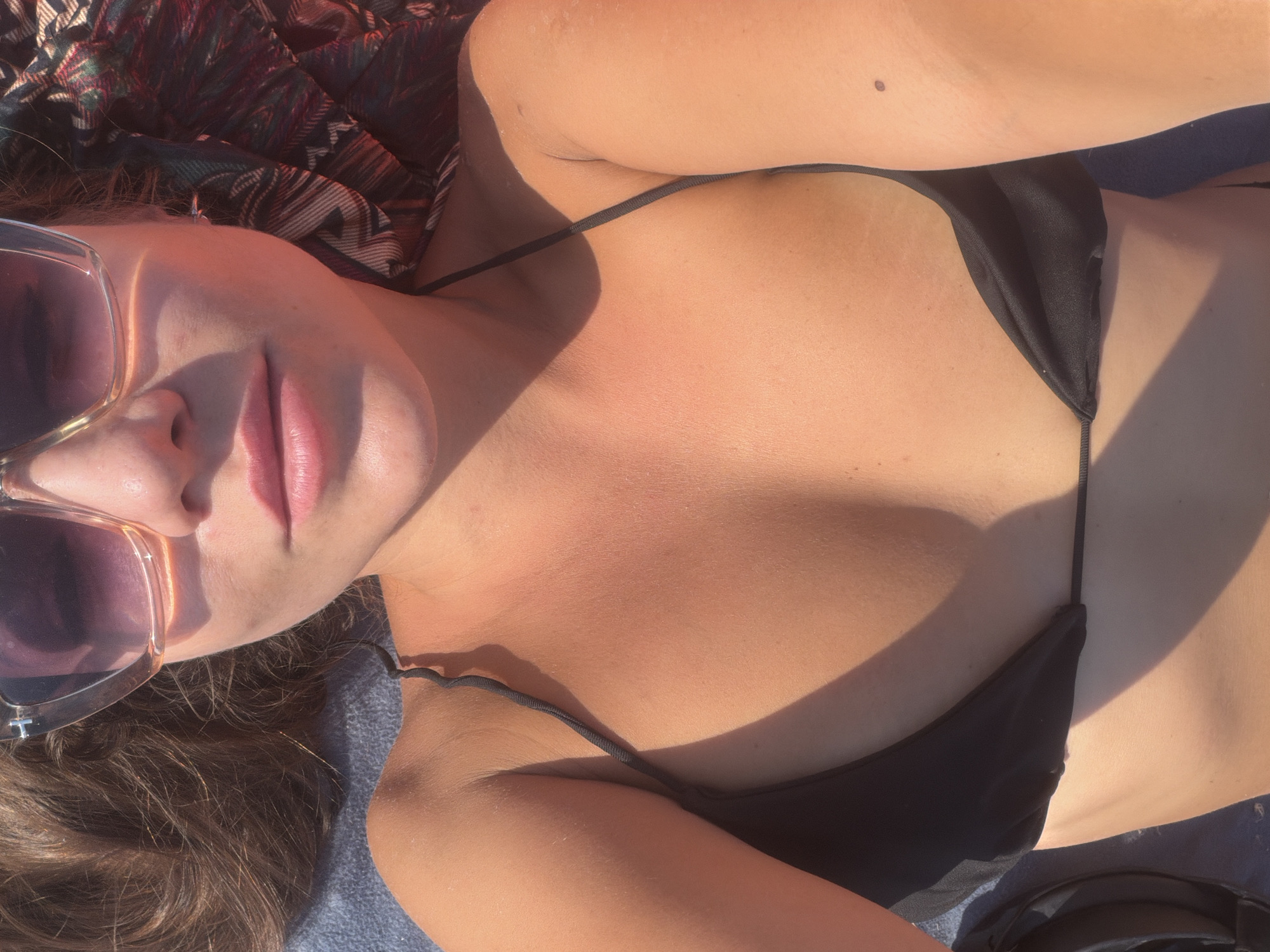 The media file is a close-up, high-angle shot of a person lying down, likely outdoors. The subject is wearing a black bikini. Sunglasses with pinkish lenses are positioned on the left side of the frame, partially obscuring the face. The person's lips are visible and appear to be slightly parted. The skin is tanned and shows highlights from sunlight, suggesting a sunny time of day. A patterned cloth, possibly a towel or sarong, is visible in the upper left corner. The background appears to be a textured surface, possibly a beach towel or blanket. The overall lighting and coloration suggest the image was taken during daylight hours, possibly late afternoon due to the warm tones and shadows. The context of "Đurovići, Montenegro" indicates the geographical location. The scene evokes a relaxed, vacation-like atmosphere, typical of a beach or poolside setting. There is no visible text in the image.