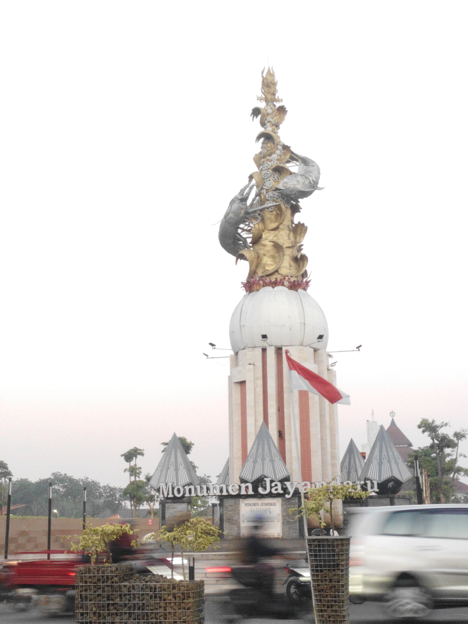 This is a medium-shot, eye-level, outdoor, daytime, vertical, slightly blurry photograph of the Jayandaru Monument in Sidoarjo, Indonesia. The monument is a tall, elaborate structure with a golden sculpture of mythical creatures at the top. A red and white Indonesian flag is attached to the side of the monument. In the foreground, there are two wire cages filled with rocks and plants, and a red vehicle is blurred due to motion. The background features trees and several buildings, including a mosque. The overall atmosphere is hazy, likely due to the weather conditions or the time of day.