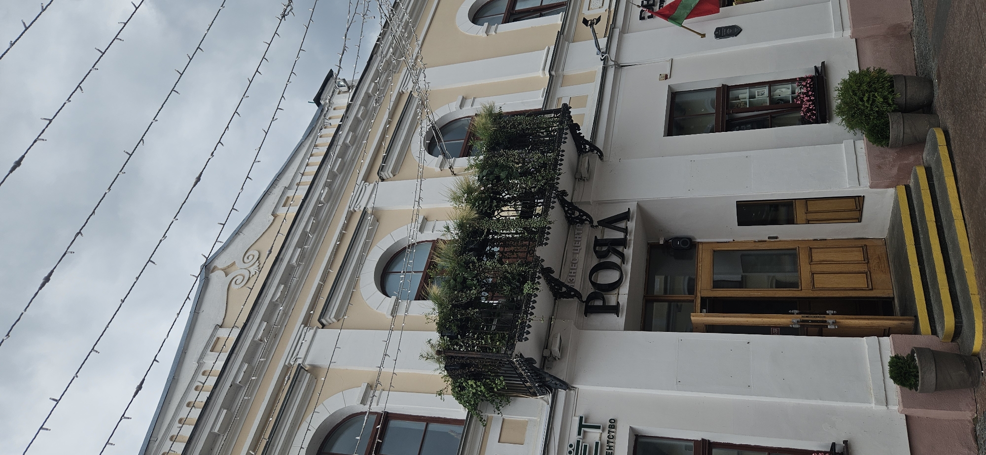 The image shows the facade of a building in Hrodna, Belarus. The building has a light yellow upper story with arched windows and decorative white molding. Below, the facade is white with a black metal balcony overflowing with greenery. A large black sign with the Cyrillic letters "РОЯЛ" is mounted above a wooden double door. The doorway has steps leading up to it, painted with yellow stripes. There are potted plants on either side of the entrance. A flag, likely the flag of Transnistria, is visible on the right side of the building. The sky is overcast. There are no people visible in the image.
