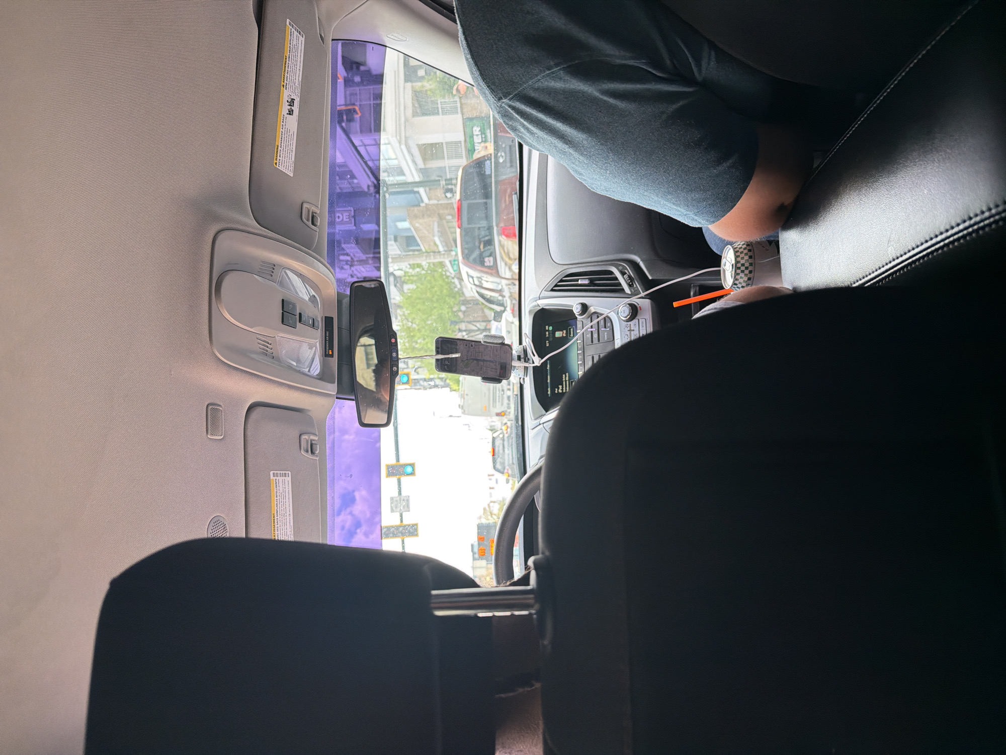 The image shows the view from the back seat of a car, looking forward through the windshield. The sun visors are down, and a smartphone is mounted on the dashboard. A person is visible in the passenger seat, wearing a blue shirt. Another person is in the driver's seat. Outside the car, the scene is a city street with buildings and traffic. The traffic lights are visible, and the buildings have awnings and storefronts. Based on the buildings in the background, it is in Atlanta, United States of America.