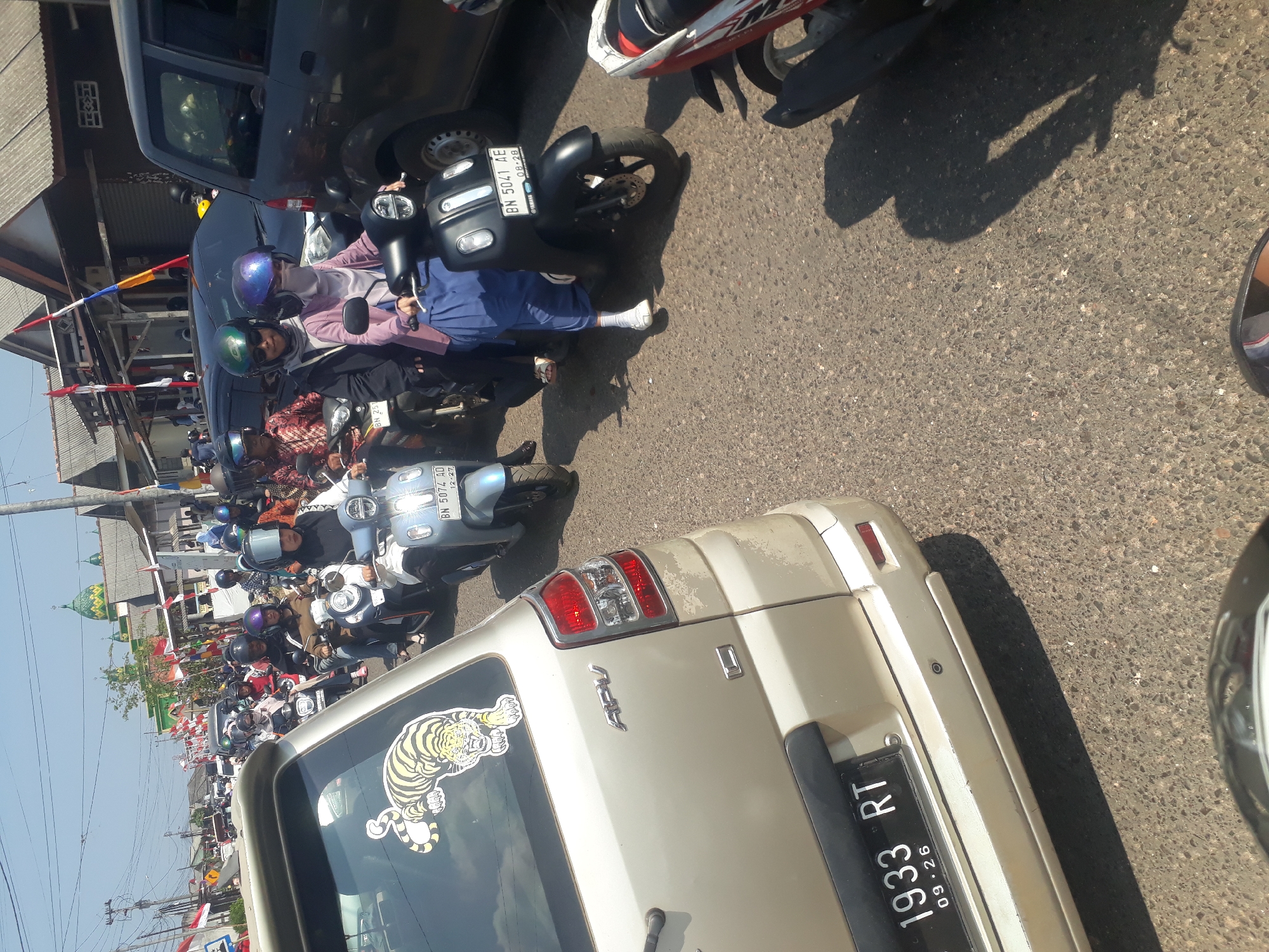 In Desa Balun Ijuk, Indonesia, on a sunny day, a traffic jam is depicted. The foreground shows the rear of a light-colored SUV, with a sticker of a tiger on its back window and its license plate reading "1933 RT 09-26". The road is filled with a line of motorcycles. On the right side, a person on a black scooter wears a purple helmet and blue pants. To the left of this scooter, a light blue scooter is ridden by a person wearing a white helmet. Further back, more motorcycles are visible, with riders wearing helmets and some covering their faces. The background reveals buildings with grey roofs and flags, suggesting a residential or commercial area. The overall scene captures a typical traffic situation in a bustling Indonesian locale.
