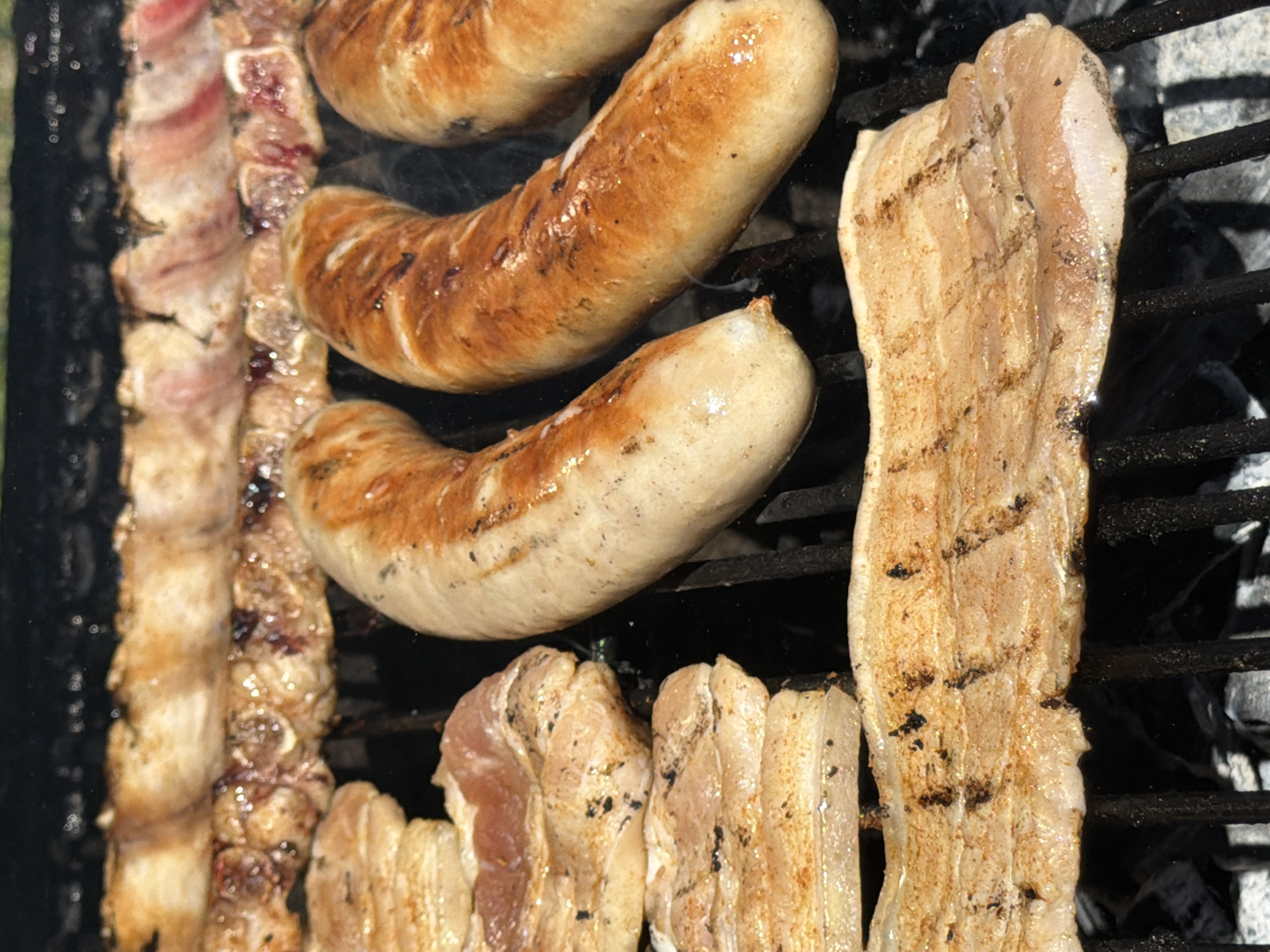 The image shows food being cooked on a grill, possibly outdoors. Visible on the grill are sausages and strips of meat, likely pork belly or bacon, grilling over heat, based on the slight browning and grill marks. The presence of a grill suggests a casual outdoor cooking scenario, perhaps a backyard barbecue. The exact location in Esch-sur-Alzette, Luxembourg, is not discernible from the image, so it is impossible to provide a specific description related to that location.