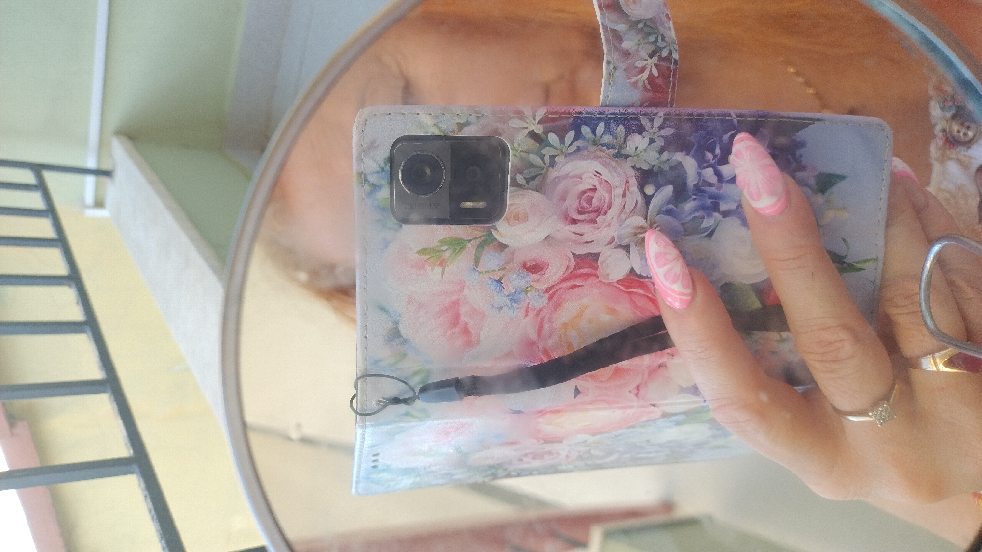 A woman's hand, adorned with pink and white marbled nail art, holds a floral-patterned phone case in front of a mirror. The phone case features a bouquet of pink and white roses with blue and purple accents. A black lanyard is attached to the case. A diamond ring is visible on the woman's ring finger. In the background, a railing and a yellow wall suggest an outdoor setting, possibly a balcony in Perea, Greece. The woman's face and shoulder, with reddish hair, are reflected in the mirror.