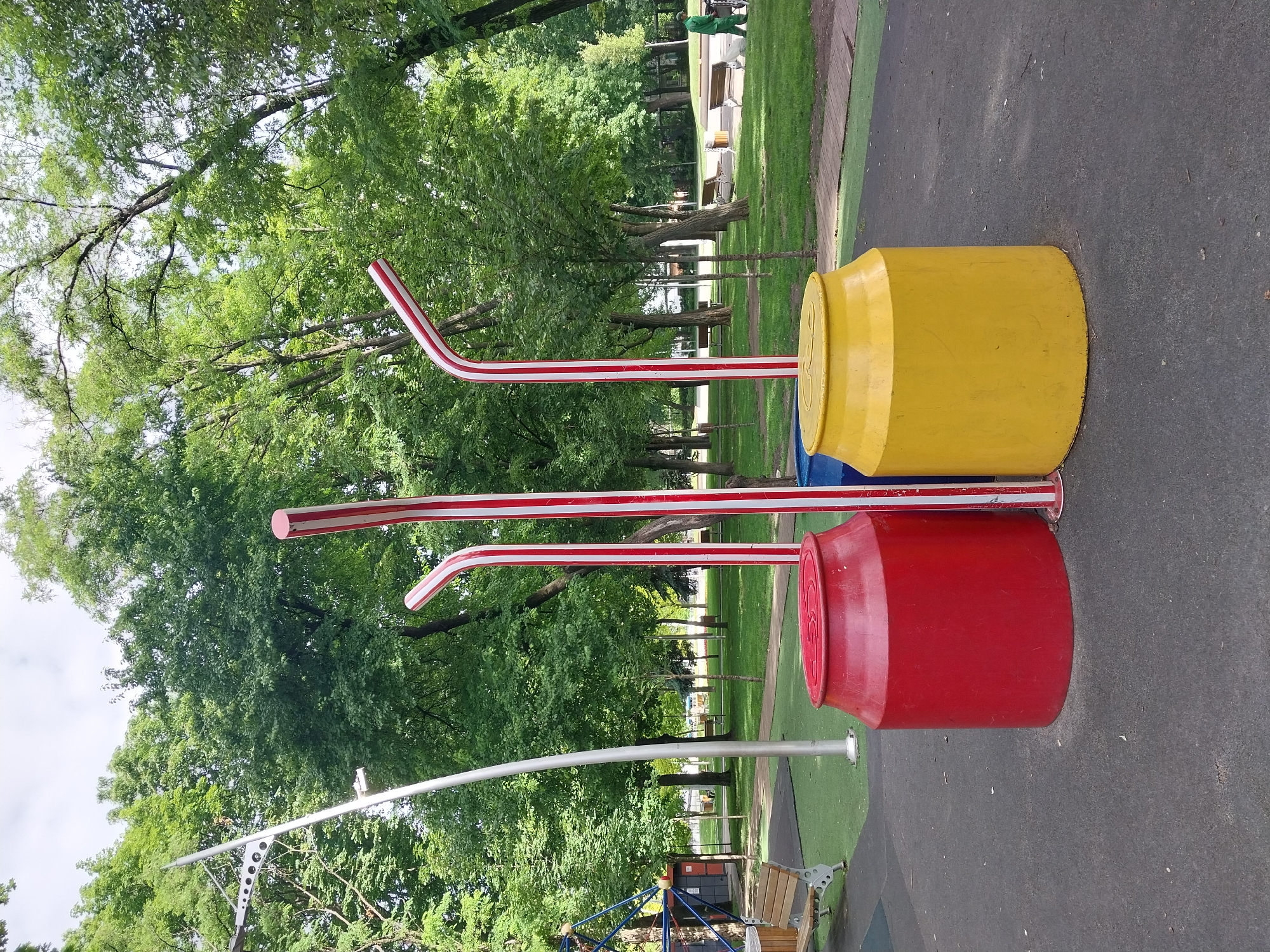 In Dnipro, Ukraine, on a sunny day, a children's playground features three oversized, brightly colored elements resembling soda cans with straws. A large red can sits to the left, a blue can is partially visible in the center, and a yellow can is to the right. Each can is topped with a giant, red-and-white striped straw. The playground has a soft, dark grey surface in the foreground, transitioning to a green synthetic turf area with benches and trees in the background. A person in a green uniform is walking in the distance on the right side of the image. The overall scene depicts a whimsical and playful outdoor recreational space.