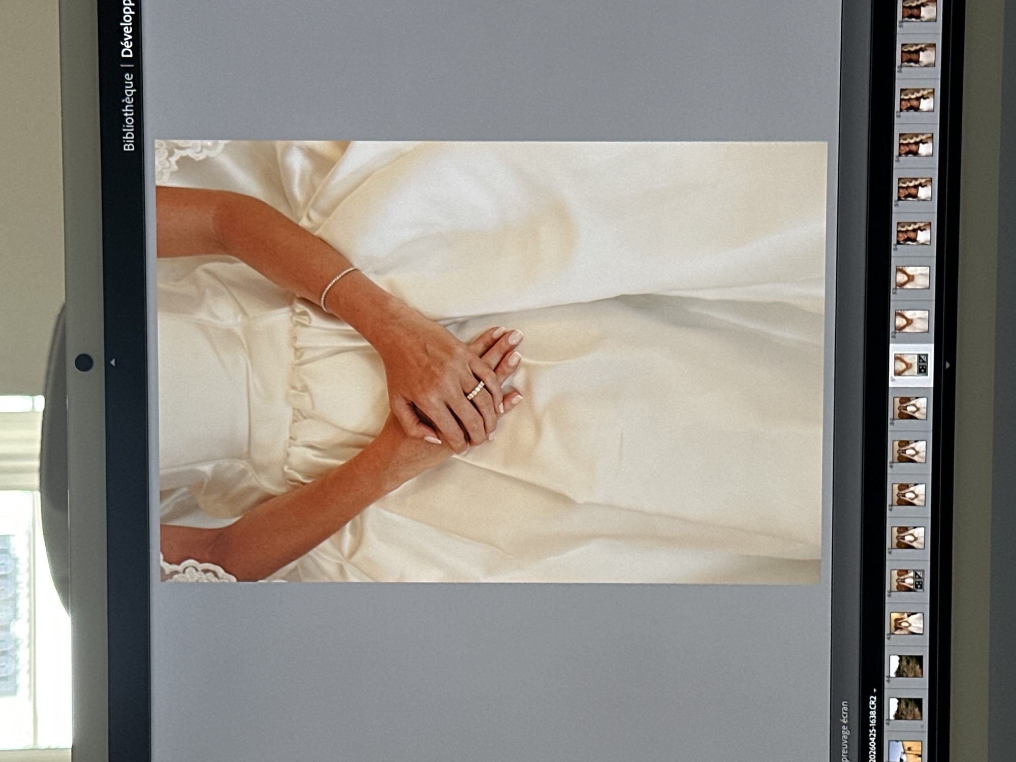 The media file displays a close-up photograph of a bride's hands resting on her white or off-white wedding dress, shown on a digital screen. The person's left hand, adorned with a multi-band wedding ring, gently holds the right hand, which wears a delicate, sparkling bracelet. The dress fabric is satin-like, with soft folds visible. The photograph is presented within a photo editing interface, likely Adobe Lightroom, indicated by the French text "Bibliothèque | Développp" (Library | Develop) on the left sidebar.
On the right side of the screen, a vertical filmstrip displays a series of other photo thumbnails, primarily showing close-ups of similar wedding-related details, some featuring parts of a wedding dress, flowers, or hands. At the bottom of this sidebar, partial text "préauvage écran" (possibly "préréglage écran" or "prévisualisation écran" - screen preview/preset) and a filename "20200425-1638.CR2" are visible, indicating the image is a RAW file captured on April 25, 2020. The overall scene suggests a professional viewing or editing session of wedding photographs. While the location context is given as Paris, France, no explicit Parisian landmarks are visible within the image itself, though the French text on the screen aligns with this context. The image captures a moment of elegance and commitment associated with a wedding.