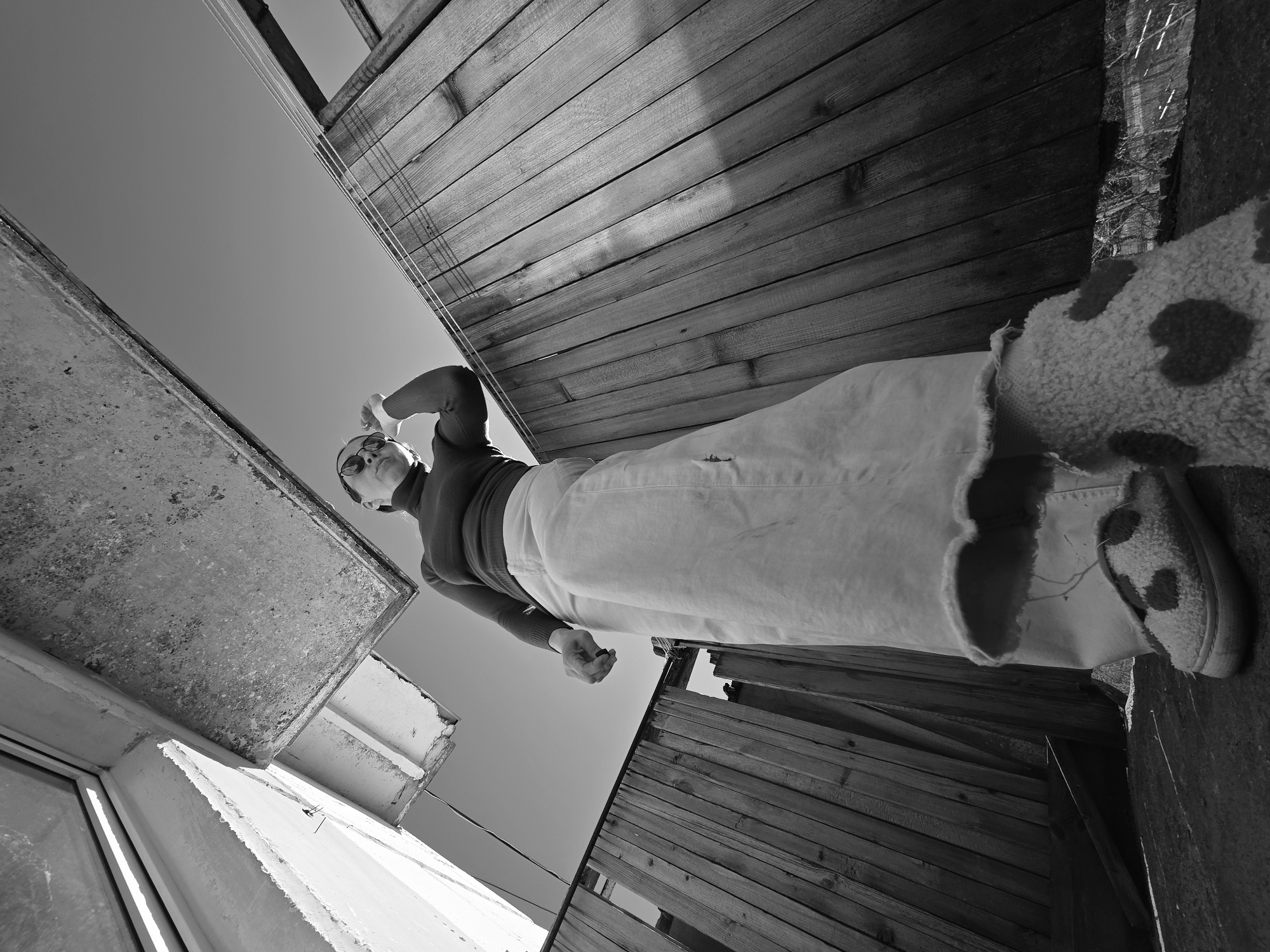 A low-angle, black and white photograph depicts a person standing between two structures. The person wears a dark, long-sleeved top, light-colored pants, and spotted, fur-lined slippers. They wear sunglasses and appear to be holding an object in one hand. Above the person, the sky is visible between the structures. One structure is a weathered, wooden fence, and the other is a building with a window and concrete overhang. The location is potentially Chaikovsky, Russia.