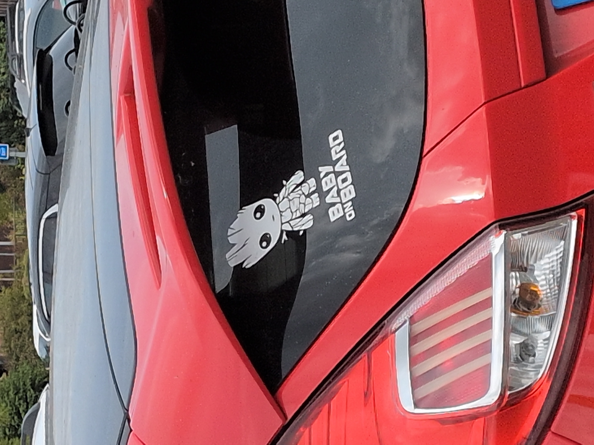A close-up, rear-view, eye-level shot shows the bumper and taillight of a red car. On the rear window of the car, a white decal of Groot from Guardians of the Galaxy is visible, with the words "BABY ON BOARD" written below it. The car is parked on a street in Telford, United Kingdom. In the background, other cars and trees are visible. The lighting suggests it is daytime.
