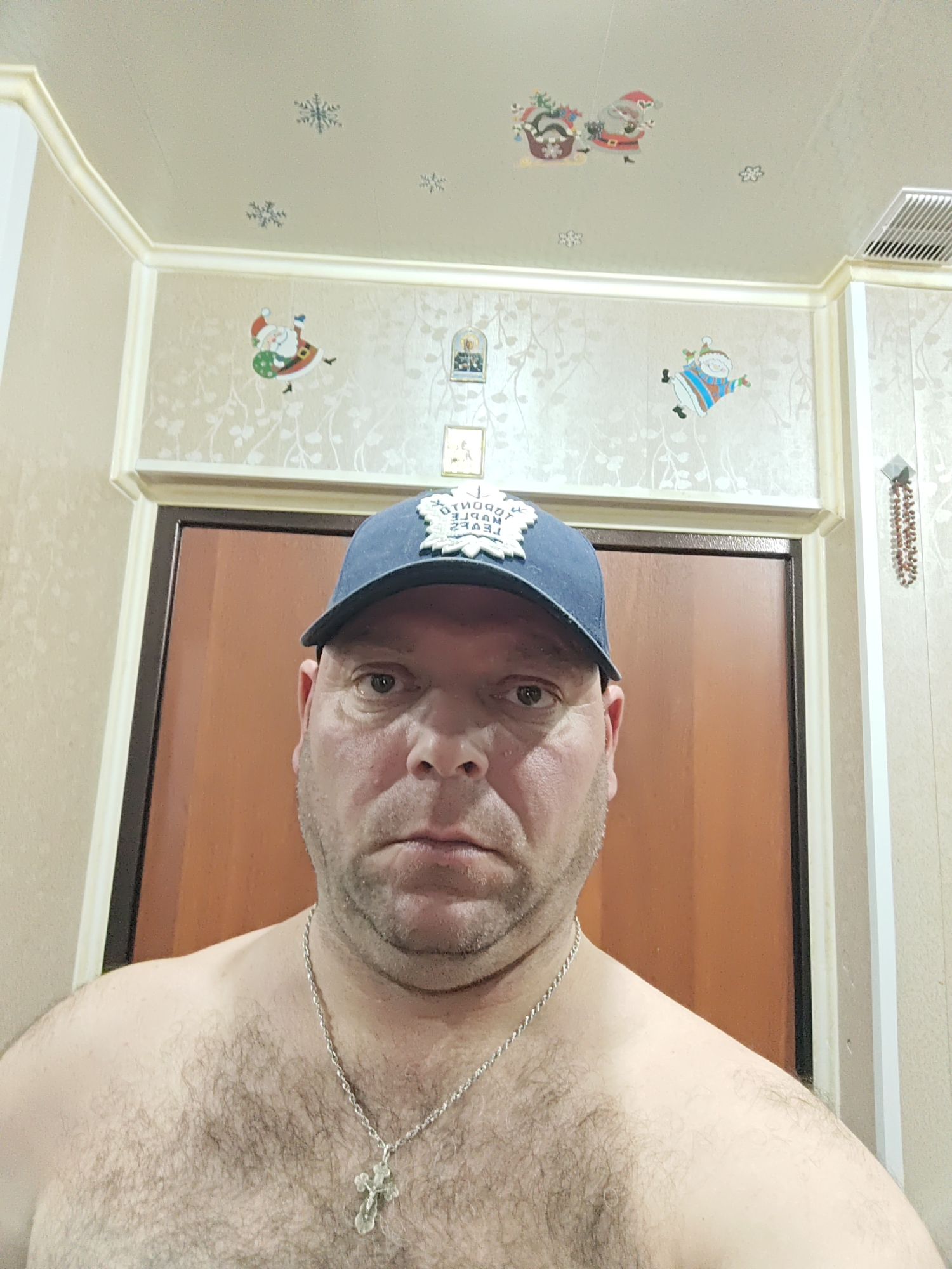 A man wearing a blue baseball cap and a silver chain with a cross necklace is posing for a selfie in what appears to be a bathroom. The cap has an emblem on the front. He has a beard and is shirtless, displaying visible chest hair. The background shows a wooden door and wallpaper with a floral pattern. There are also Christmas-themed decorations, including snowflakes and images of Santa Claus, on the ceiling and walls, suggesting a festive or holiday setting. The man's expression is neutral. The location is stated as the city of Polyarny, Russia.