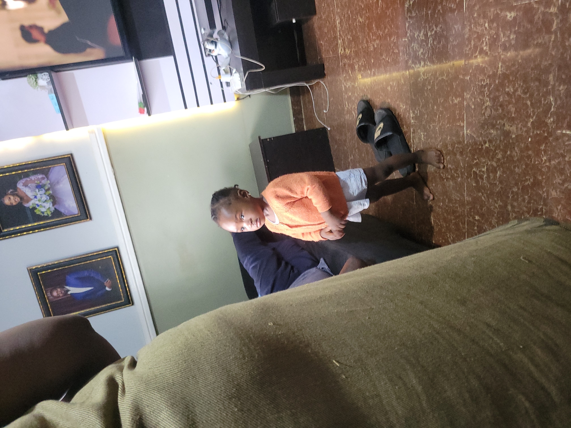 A child is standing near a person sitting on a couch in what appears to be an indoor living space. The child is wearing an orange sweater and light-colored shorts. The person on the couch is wearing a dark blue shirt and appears to be wearing shorts. The couch is green. There are two framed pictures on the wall, along with a TV and various pieces of furniture in the background. A pair of sandals is on the floor. The location is Unknown, Unknown.