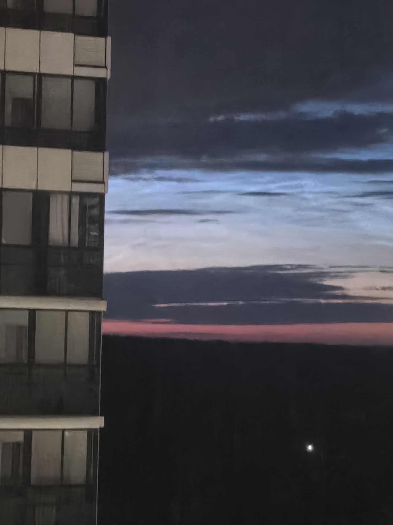 An exterior, vertical, medium shot captures a tall, multi-story building in the foreground, its facade composed of alternating dark and light rectangular sections, indicative of windows and structural elements. To the right, the sky transitions through various shades of twilight. A deep, dark blue band of clouds occupies the upper portion, followed by thinner, lighter blue and white striated clouds, suggesting atmospheric diffusion of light. Lower still, a band of dark grey clouds sits above a horizon line rendered in a vivid pinkish-red hue, indicative of sunset or sunrise. Below this colorful horizon, the landscape is a dark, indistinct mass, dotted with what appears to be a single, bright artificial light source in the distance. The scene is devoid of any visible people or animals.