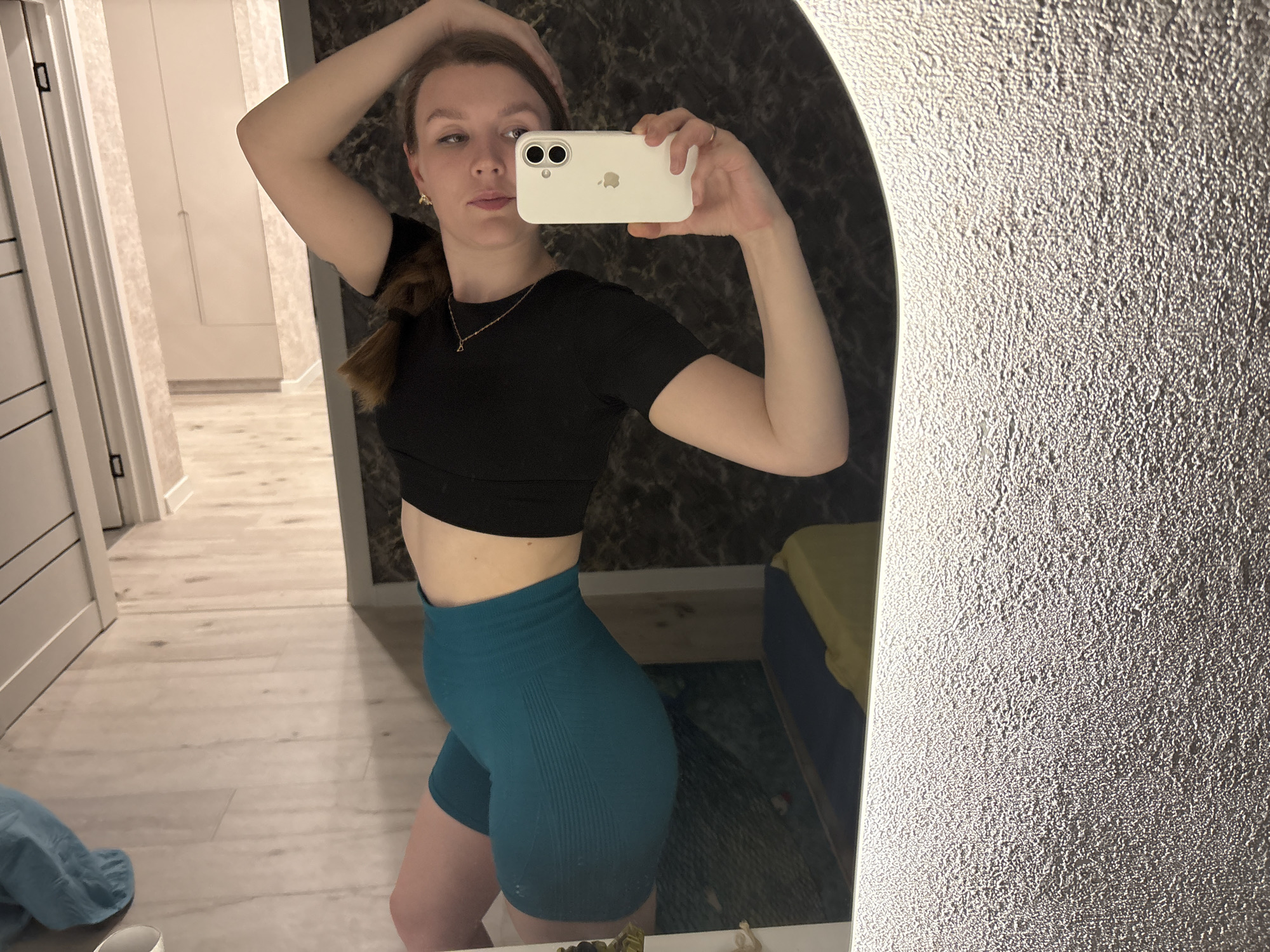 A young woman is taking a mirror selfie in what appears to be a home interior. She is wearing a black crop top and teal shorts. She holds a white smartphone in her right hand, using it to capture the image. Her left hand is raised, touching her head. A hallway is visible in the background, with a closed door and a light-colored floor. The mirror's frame and wall texture are visible on the right side of the image, and part of a bed or couch is visible in the reflection. The location cannot be ascertained as Izhevsk based on the image.